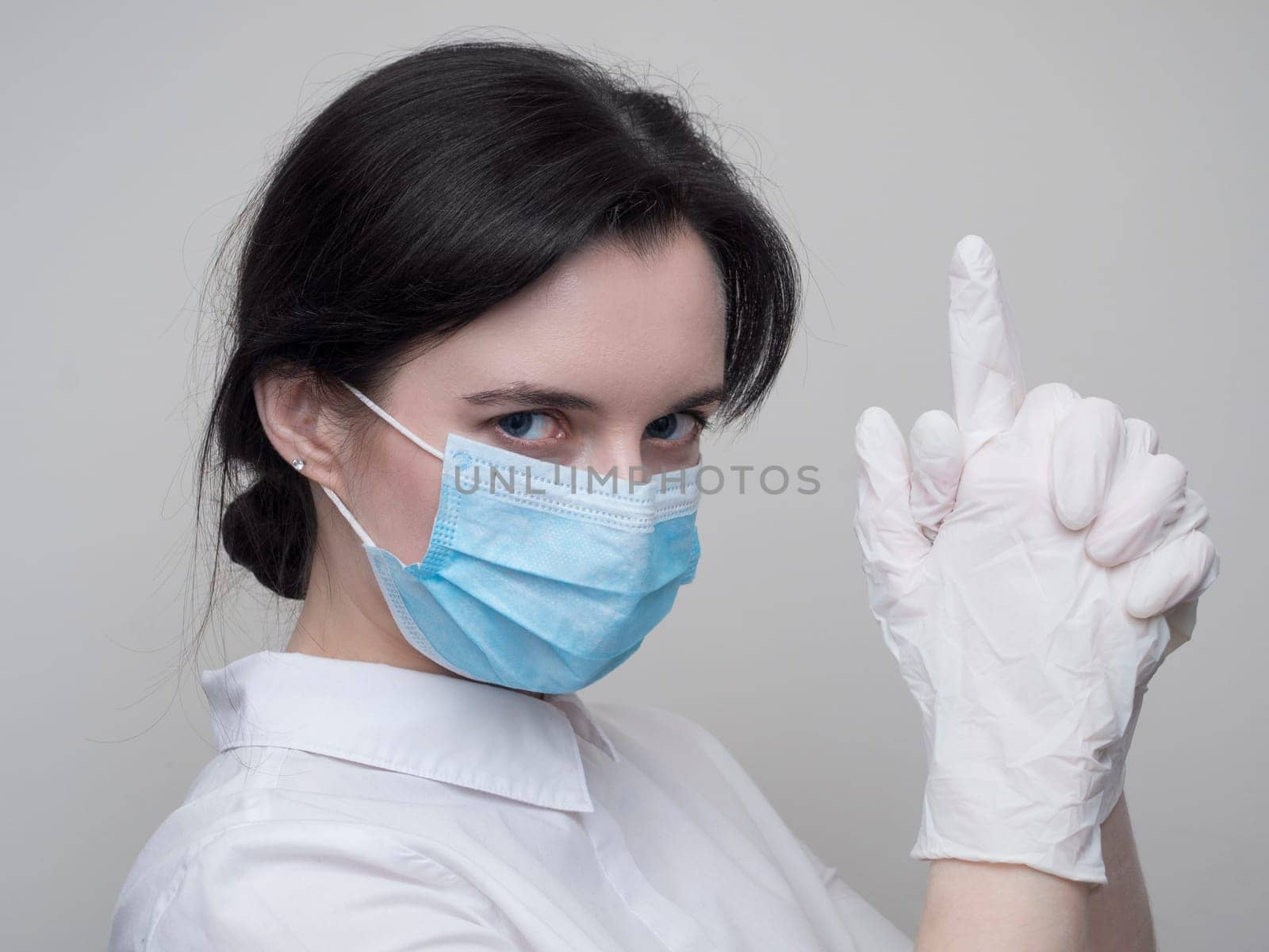 Young woman patient in a medical mask puts on protective surgical sterile gloves on her arm, on gray background, protection against coronovirus