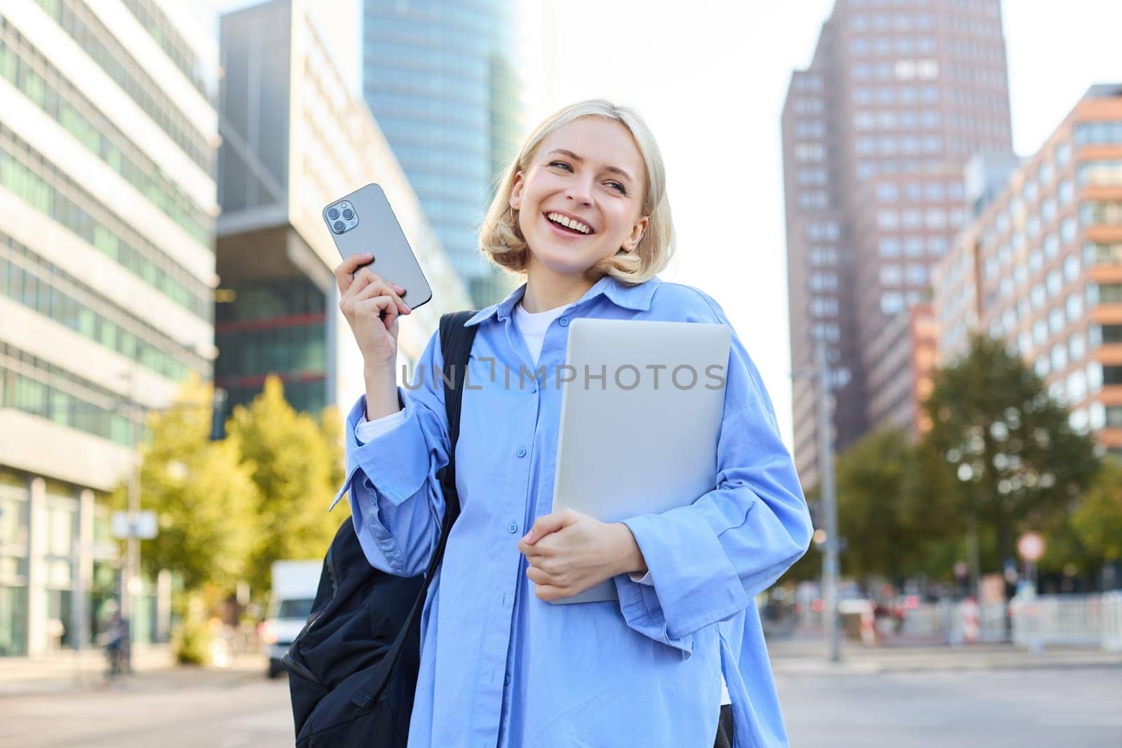 Image of young stylish, modern blond woman in blue shirt, holding laptop and backpack, using smartphone, smiling, walking along the street, posing in city centre.