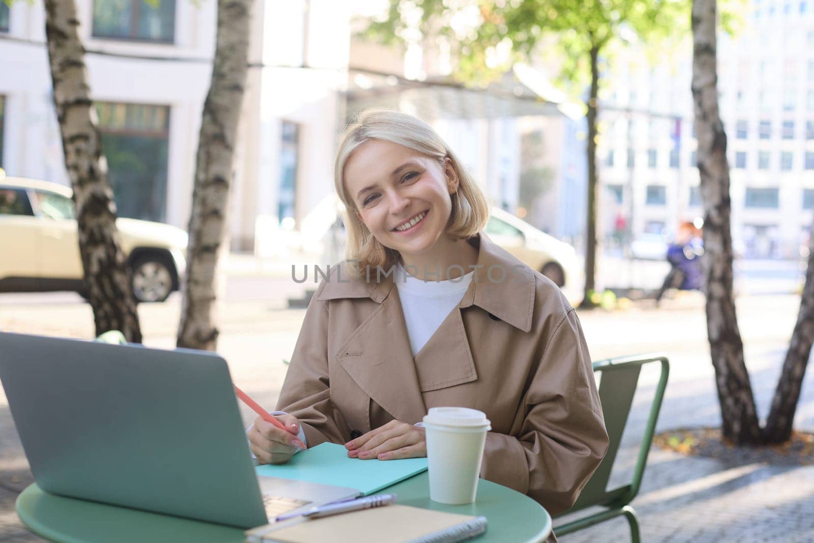 Remote workplace. Smiling young woman freelancer, student using laptop in outdoor cafe, talking to someone, attends online lecture, drinking coffee by Benzoix