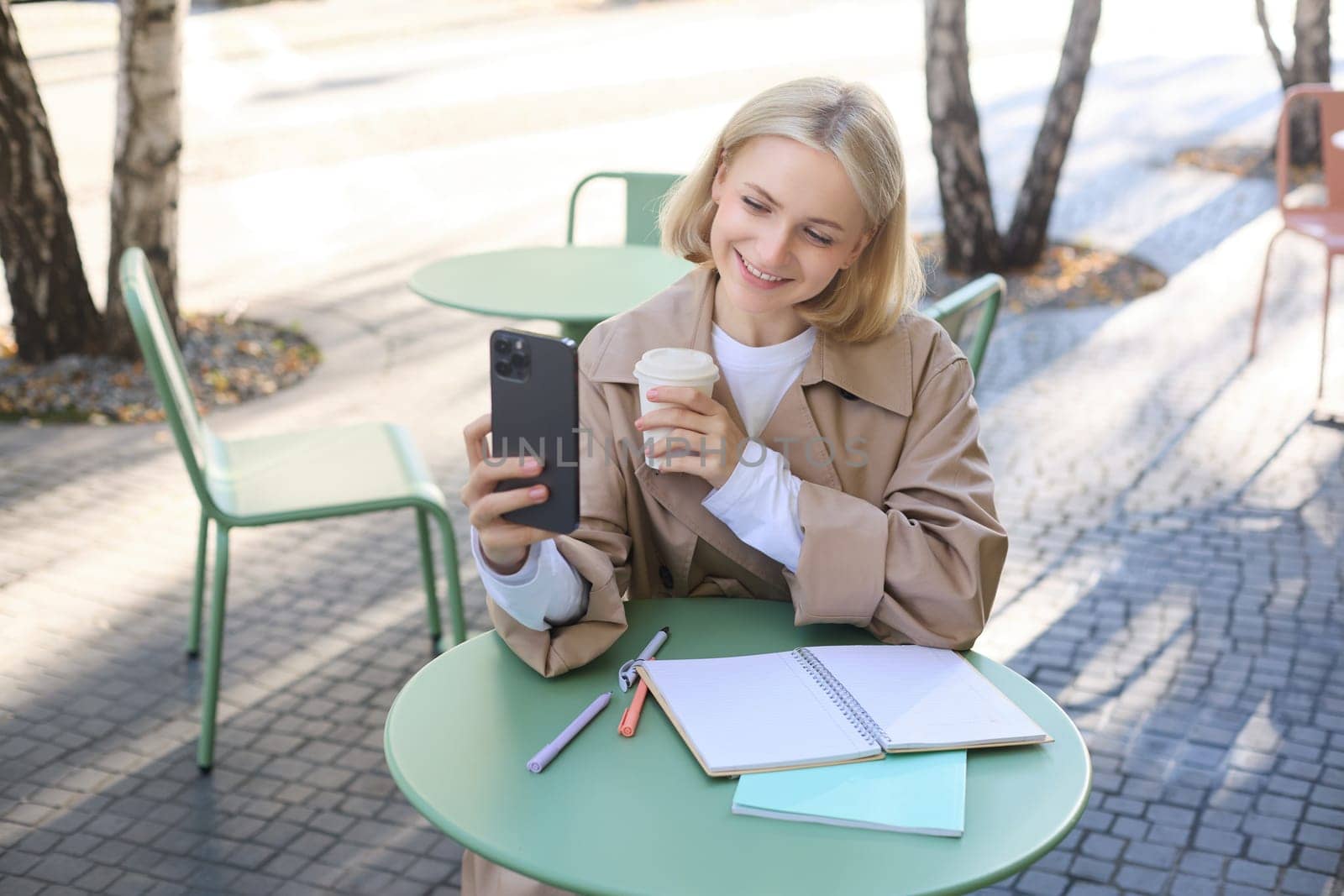 Image of stylish young woman, student taking selfie in cafe on street, posing with her favourite drink, enjoying coffee and making content for social media blog by Benzoix