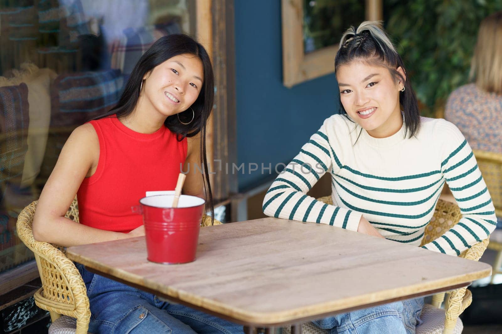 Smiling young Asian girlfriends in casual clothing sitting together at wooden table and discussing plans while spending time in cafe