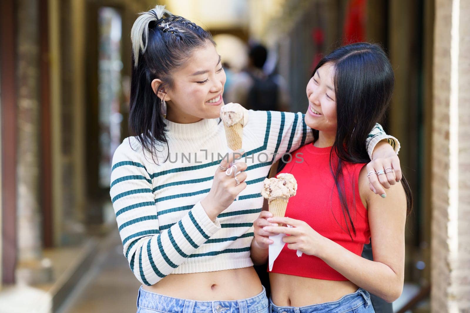 Cheerful Asian female standing with hand on shoulder of friend while eating delicious ice cream in street against blurred background