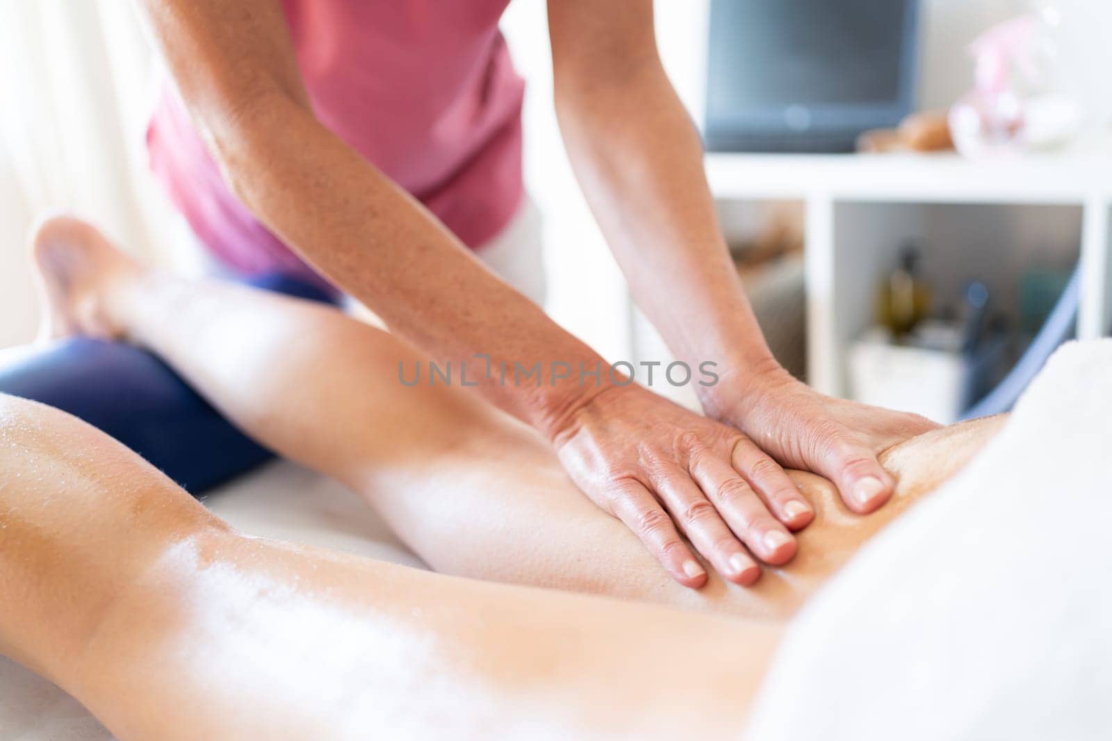 Crop female therapist rubbing oily leg of female client lying on stomach over spa table during physiotherapist massage session in clinic