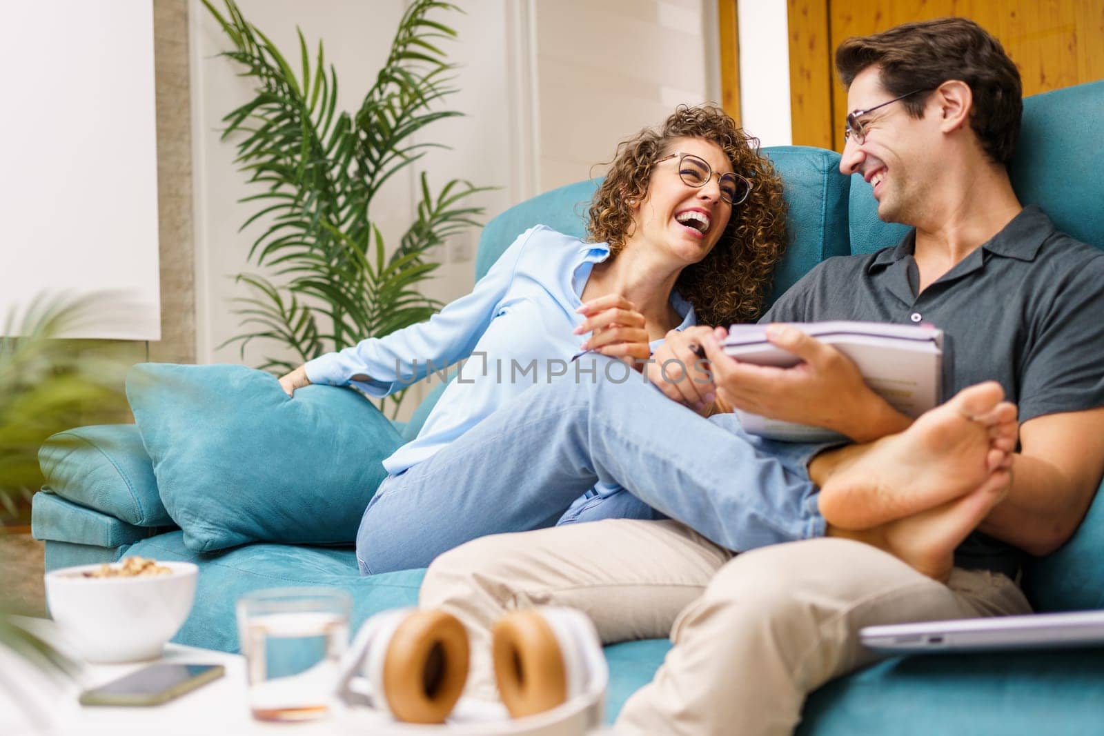 Happy young wife in casual clothes sitting with legs stretched on husband with notebook while laughing bonding together over sofa in cozy living room