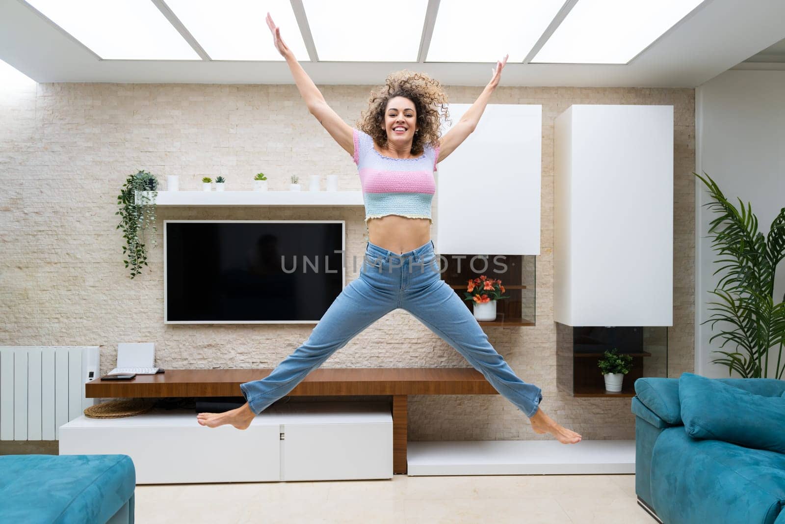 Excited young female with curly hair in casual clothes jumping above ground and raising arms while having fun at home