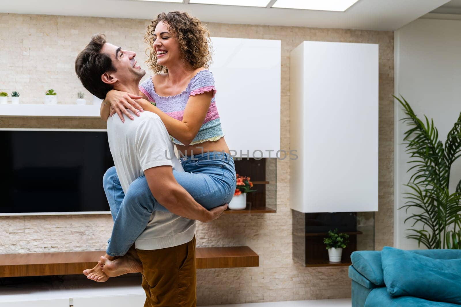 Side view of cheerful young couple hugging while standing against TV and shelves in modern apartment with potted plants on wall at home