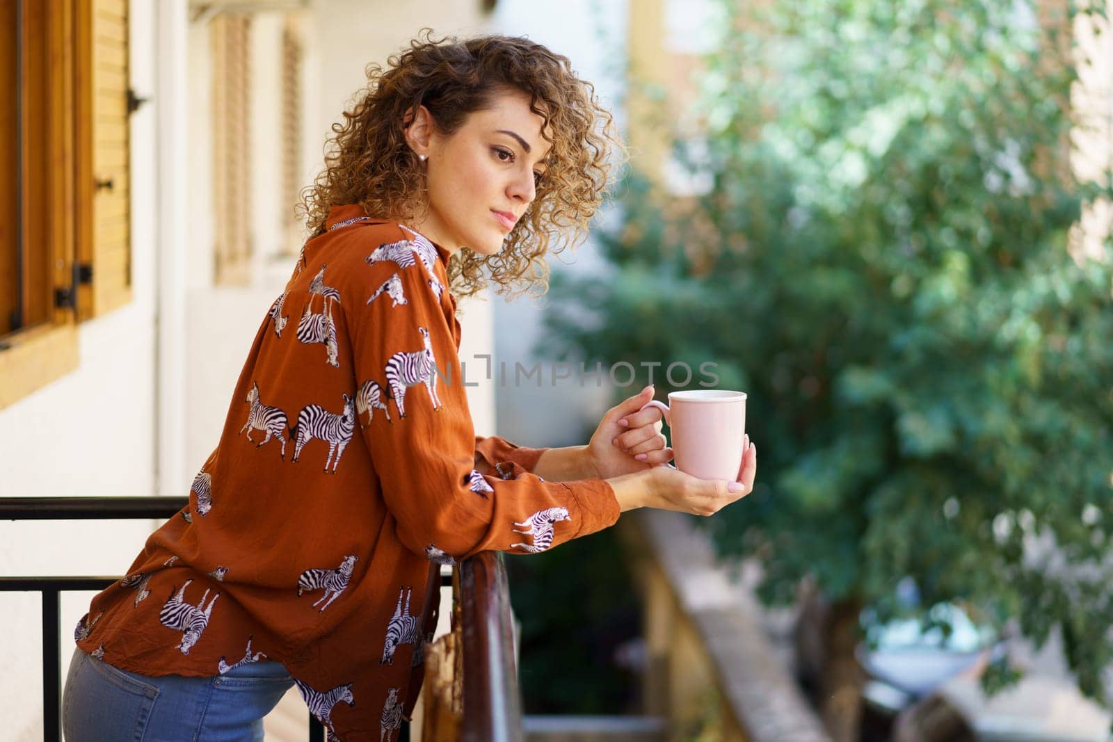 Side view of young curly haired female wearing casual clothes with coffee cup standing against blurred background in modern building
