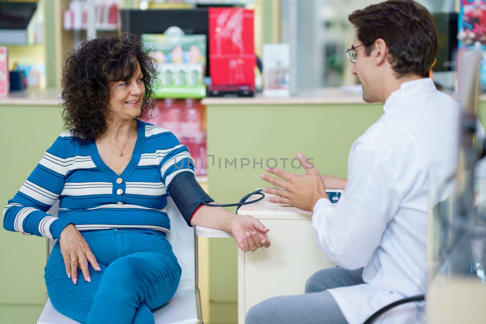 Competent pharmacist consulting female patient during blood pressure measurement while sitting together in pharmacy and looking at each other