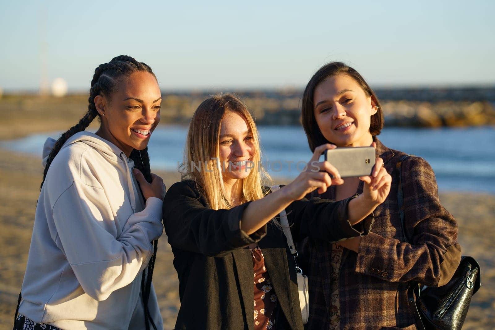 Positive multiethnic girlfriends smiling happily while taking self portrait on cellphone on sandy coast of sea at sunset