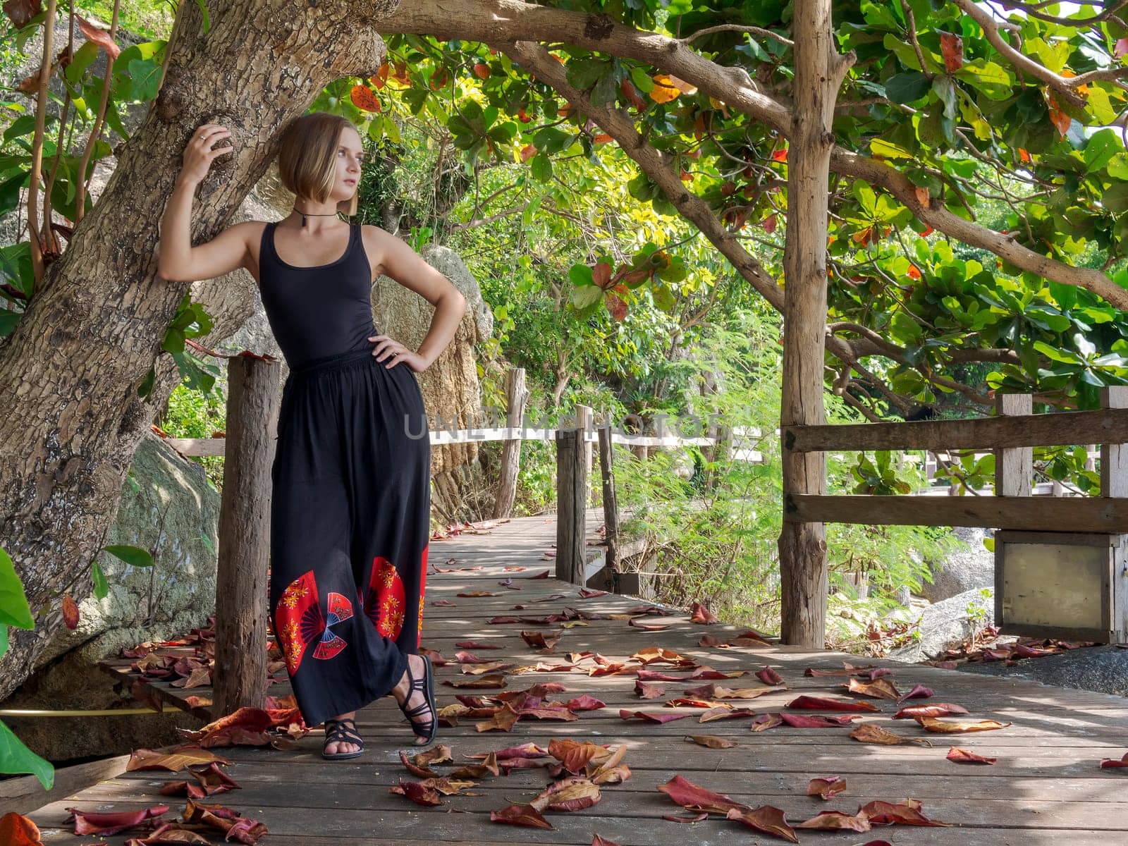 A young woman in black clothes poses by a tree in a rainforest on a wooden floor in fallen red foliage. walk in the forest on a tropical island. by Andre1ns