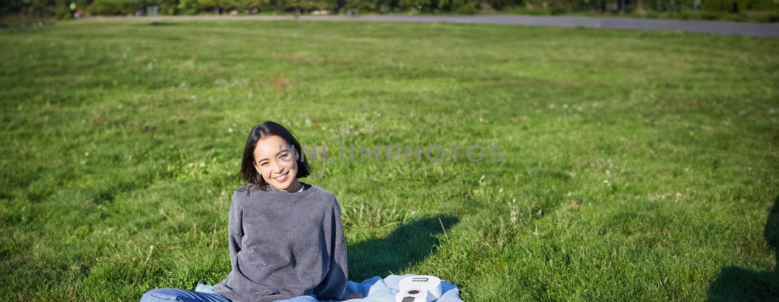 Vertical shot of smiling asian girl sitting on blanket with ukulele guitar, relaxing on sunny day outdoors, resting in park.
