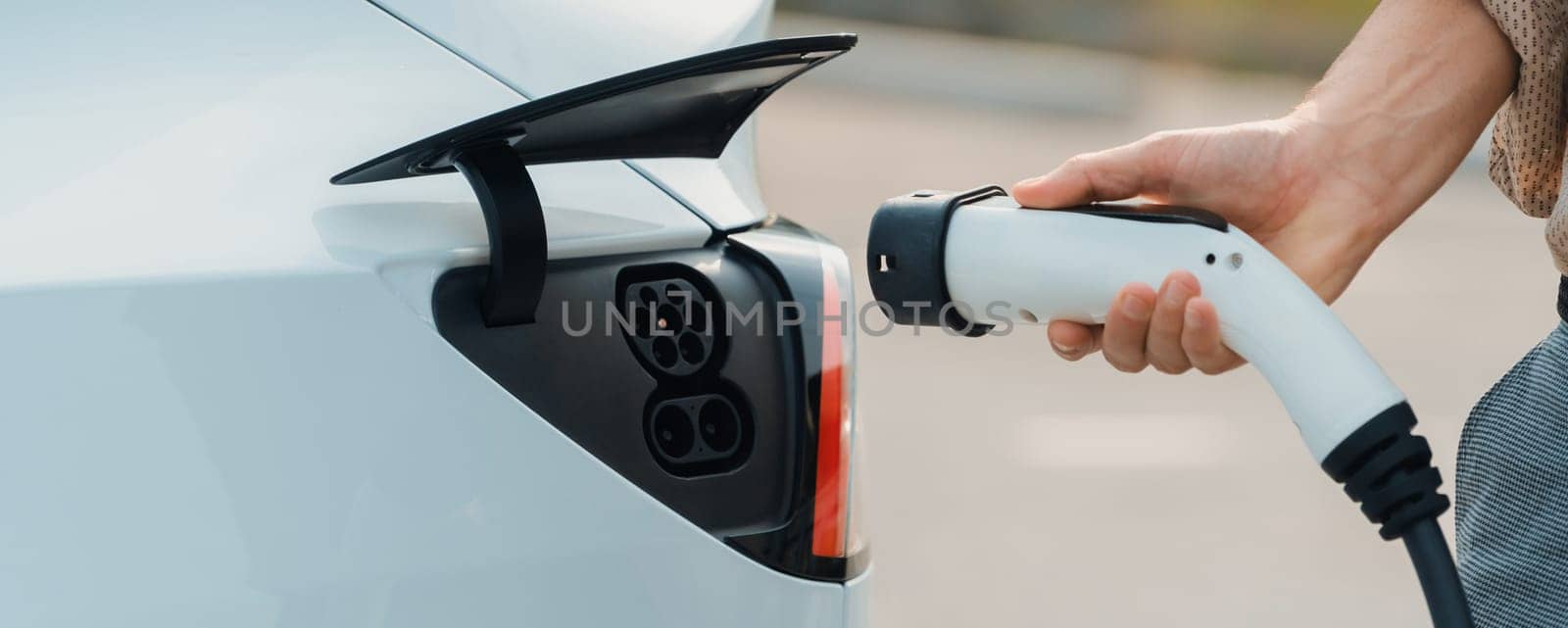 Young man recharge electric car's battery from charging station in city commercial parking lot. Rechargeable EV car for sustainable environmental friendly urban travel. Panorama Expedient