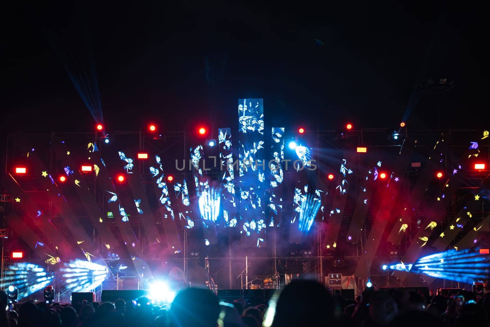 Amid vibrant nightlife concert festival takes center stage and unrecognizable crowd cheers in front of brilliantly illuminated stage. lens flare captures the energy and excitement of the music event. by Sorapop