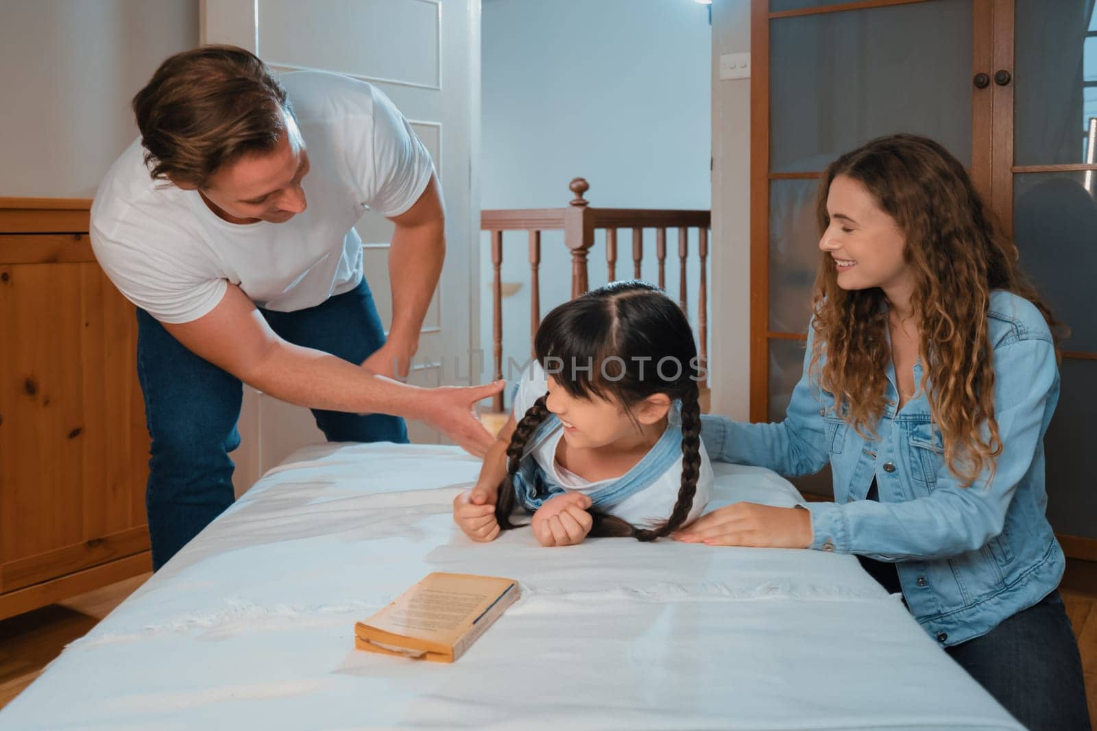 Happy modern family wakes up their little girl on the weekend with playful tickle expressing their love and affection for their young daughter, laughing and smiling together. Synchronos