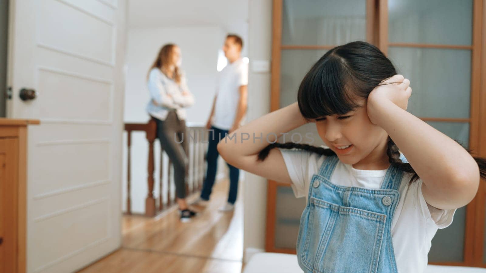 Stressed and unhappy young girl huddle in corner, cover her ears blocking sound of her parent arguing in background. Domestic violence at home and traumatic childhood develop to depression. Synchronos