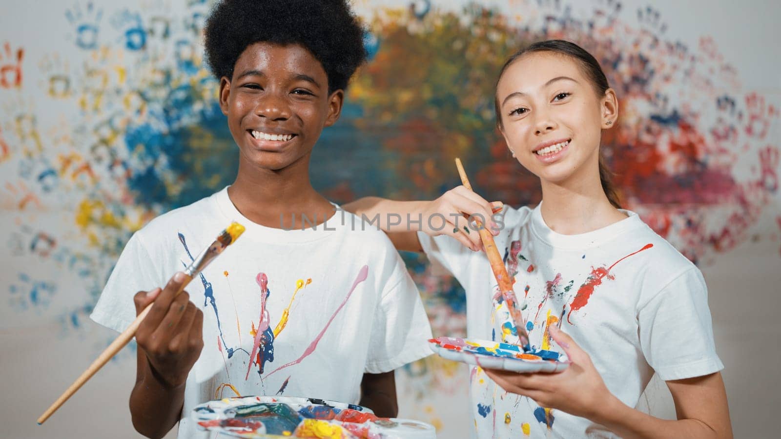 Highschool student with mixed races standing and looking at camera at art lesson. Diverse children hold the paintbrush while pose with confident at colorful stained wall. Creative style. Edification.