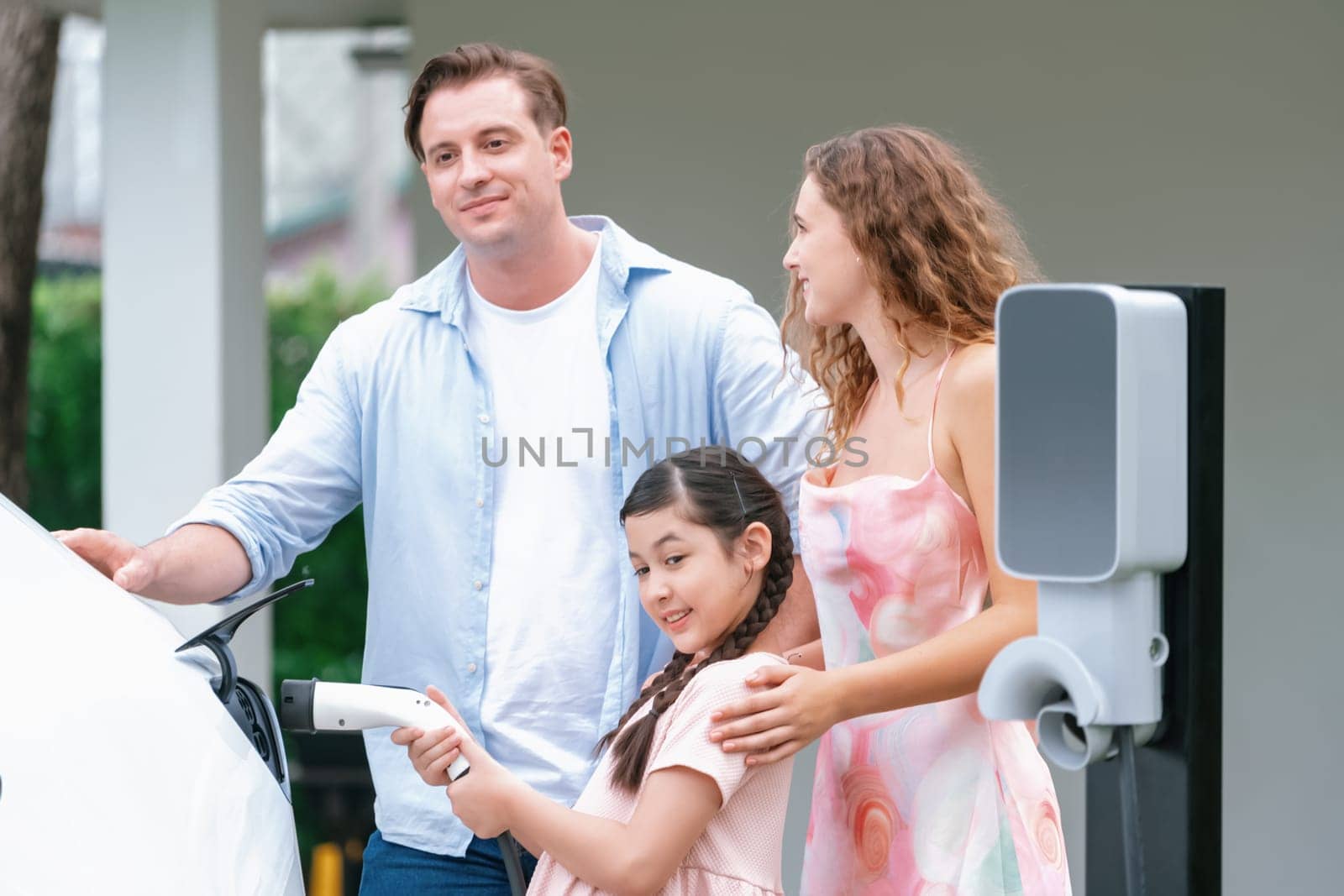 Happy little young girl learn about eco-friendly and energy sustainability as she help her family recharge electric vehicle from home EV charging station. EV car and modern family concept. Synchronos