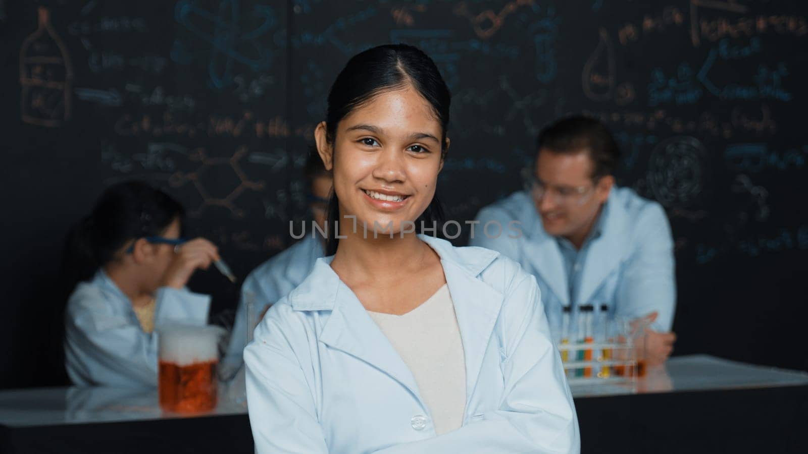 Girl looking at camera with arm folded while people doing experiment at laboratory. Cute student standing blackboard with chemical theory with blurring background at STEM science class. Edification.