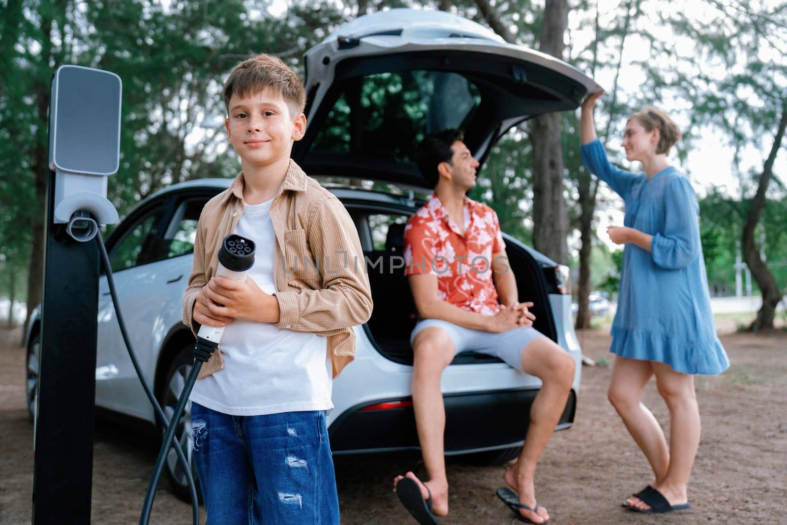 Little boy holding EV charger and point at camera with his family sitting on the trunk in background. Road trip travel with alternative energy charging station for eco-friendly car concept. Perpetual