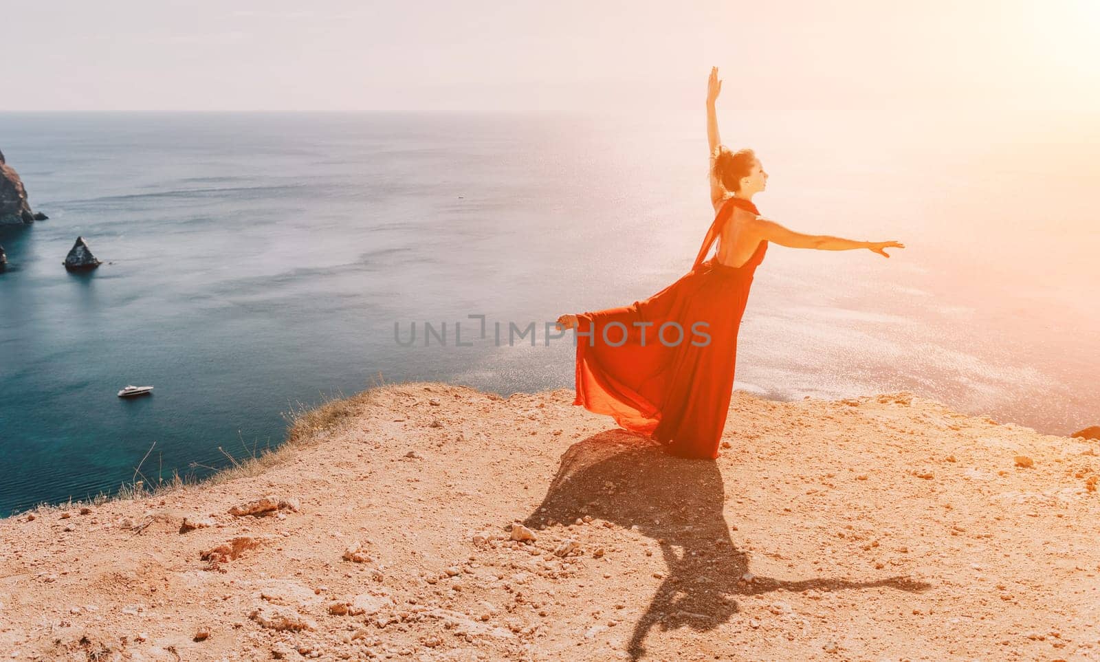 Woman in red dress on sea. Side view a Young beautiful sensual woman in a red long dress posing on a rock high above the sea on sunset. Girl on the nature on blue sky background. Fashion photo. by panophotograph