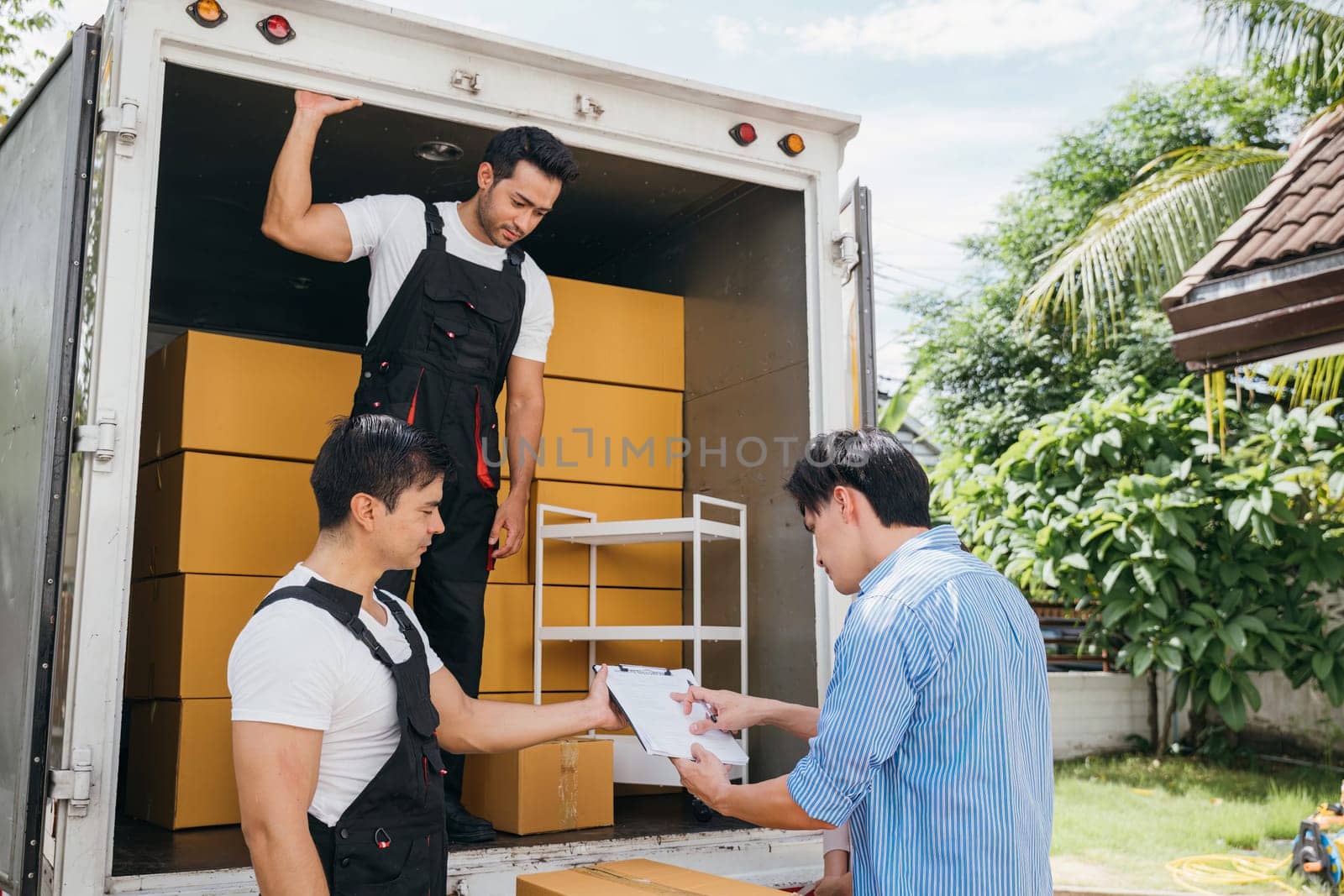 Professional movers assist a satisfied couple signing the delivery checklist after furniture lifting. Uniformed employees display teamwork for customer delight. Moving Day Concept