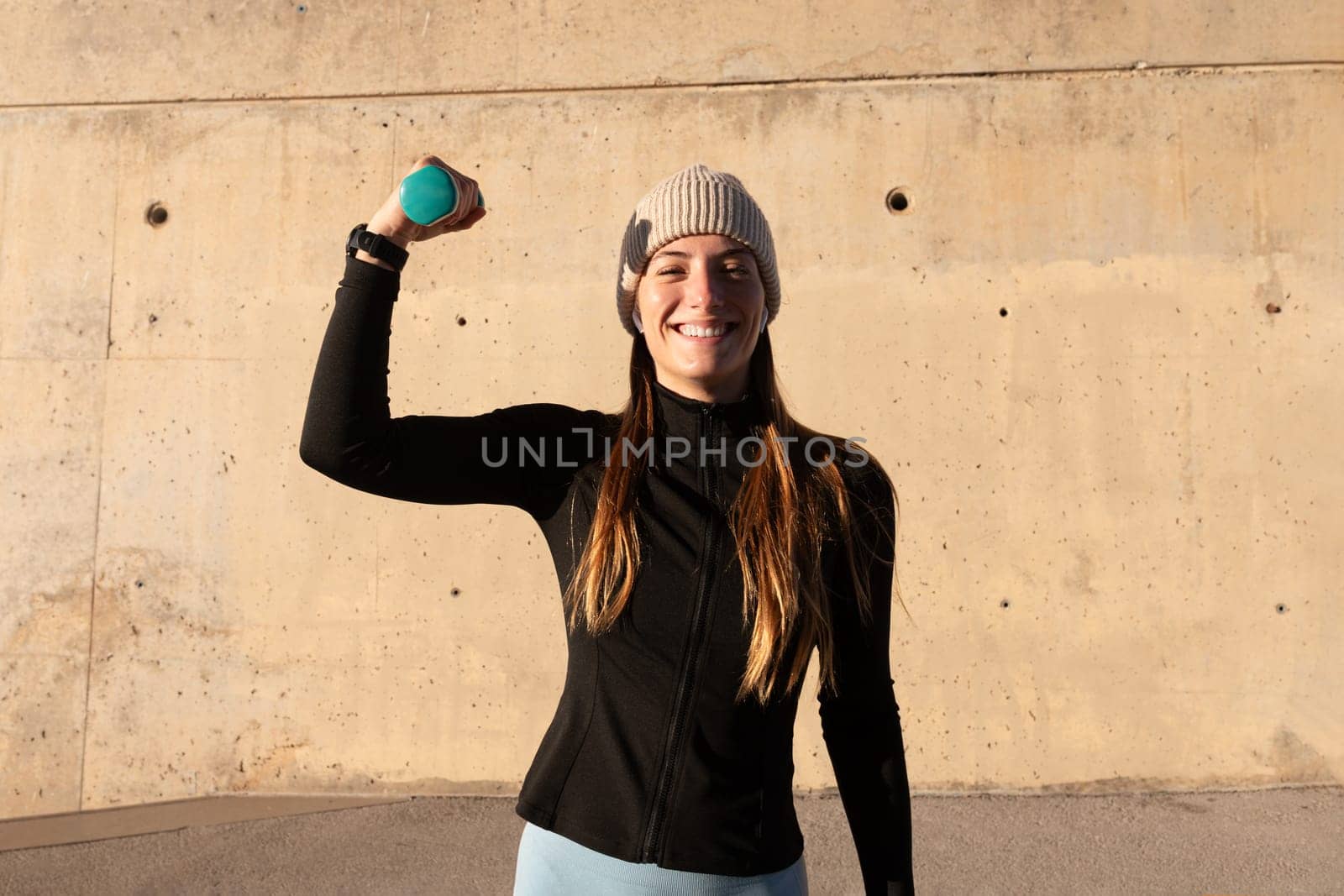 Young happy sporty woman showing strong arm holding weights looking at camera. Female showing strength holding dumbbell. by Hoverstock