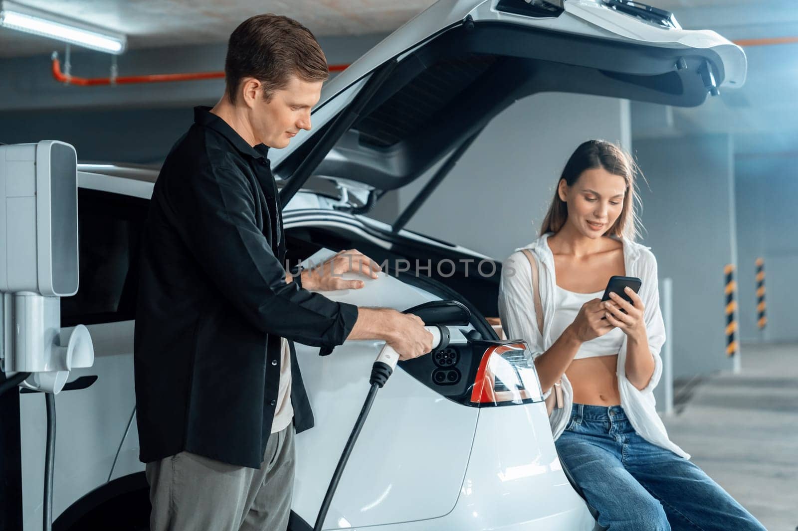 Young couple travel with EV electric car to shopping center parking lot charging in downtown city showing urban sustainability lifestyle by green clean rechargeable energy of electric vehicle innards