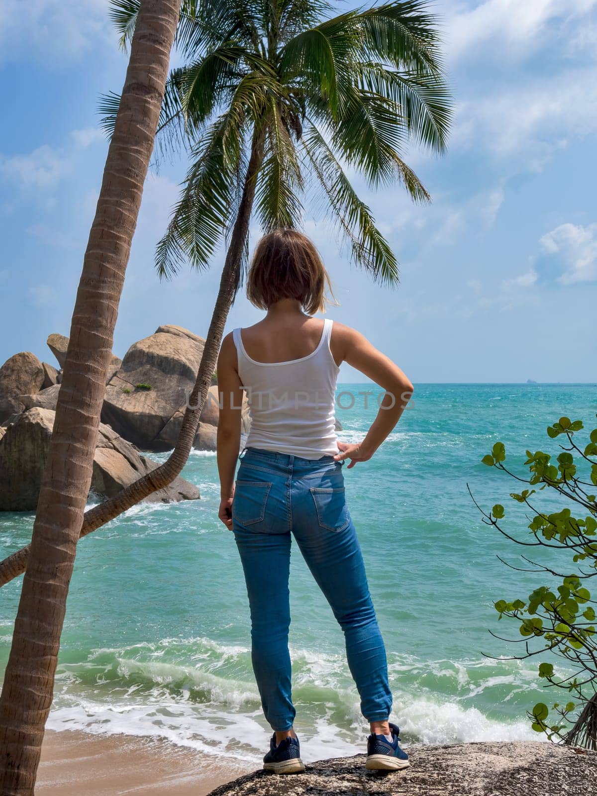 A young girl in jeans and a white T-shirt stands and looks at the sea under a palm tree. by Andre1ns