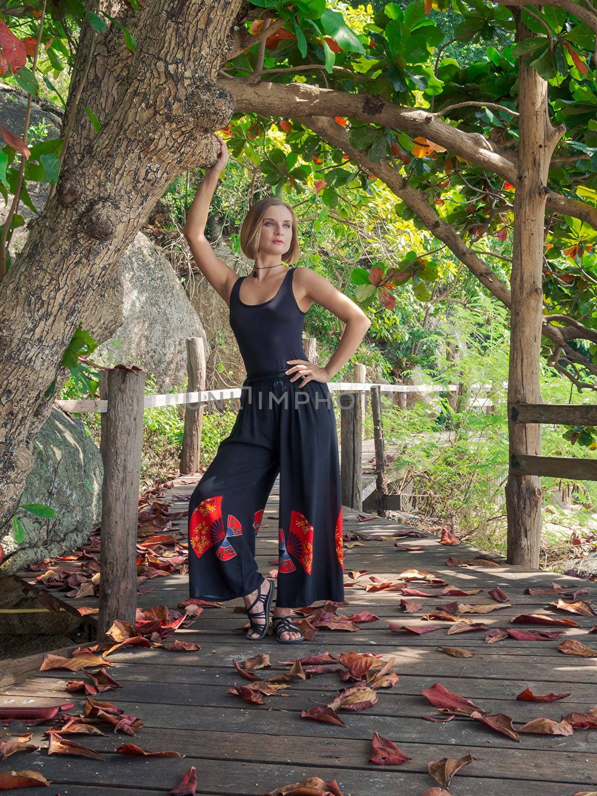 A young woman in black clothes poses by a tree in a rainforest on a wooden floor in fallen red foliage. walk in the forest on a tropical island. by Andre1ns