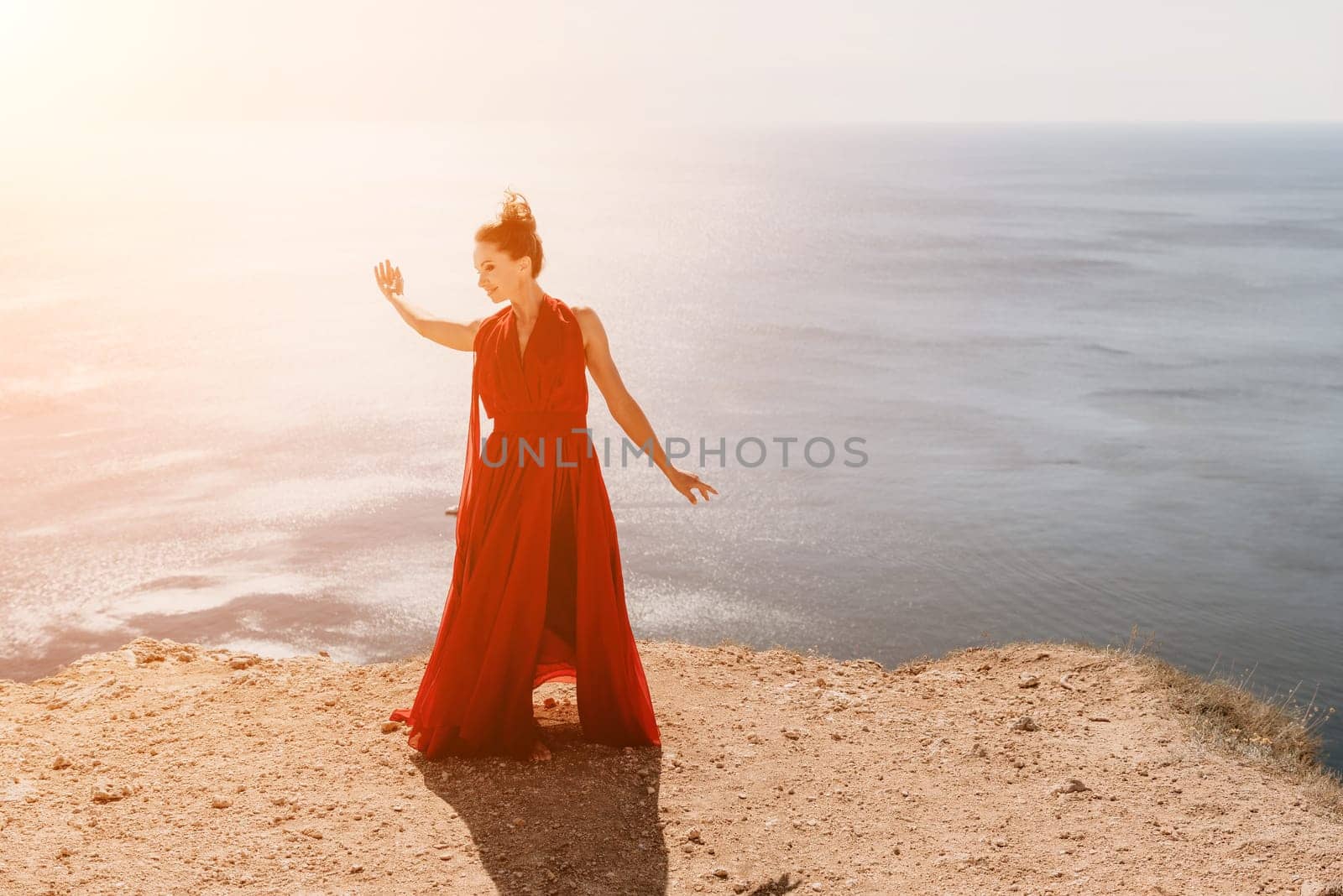 Side view a Young beautiful sensual woman in a red long dress posing on a rock high above the sea during sunrise. Girl on the nature on blue sky background. Fashion photo.