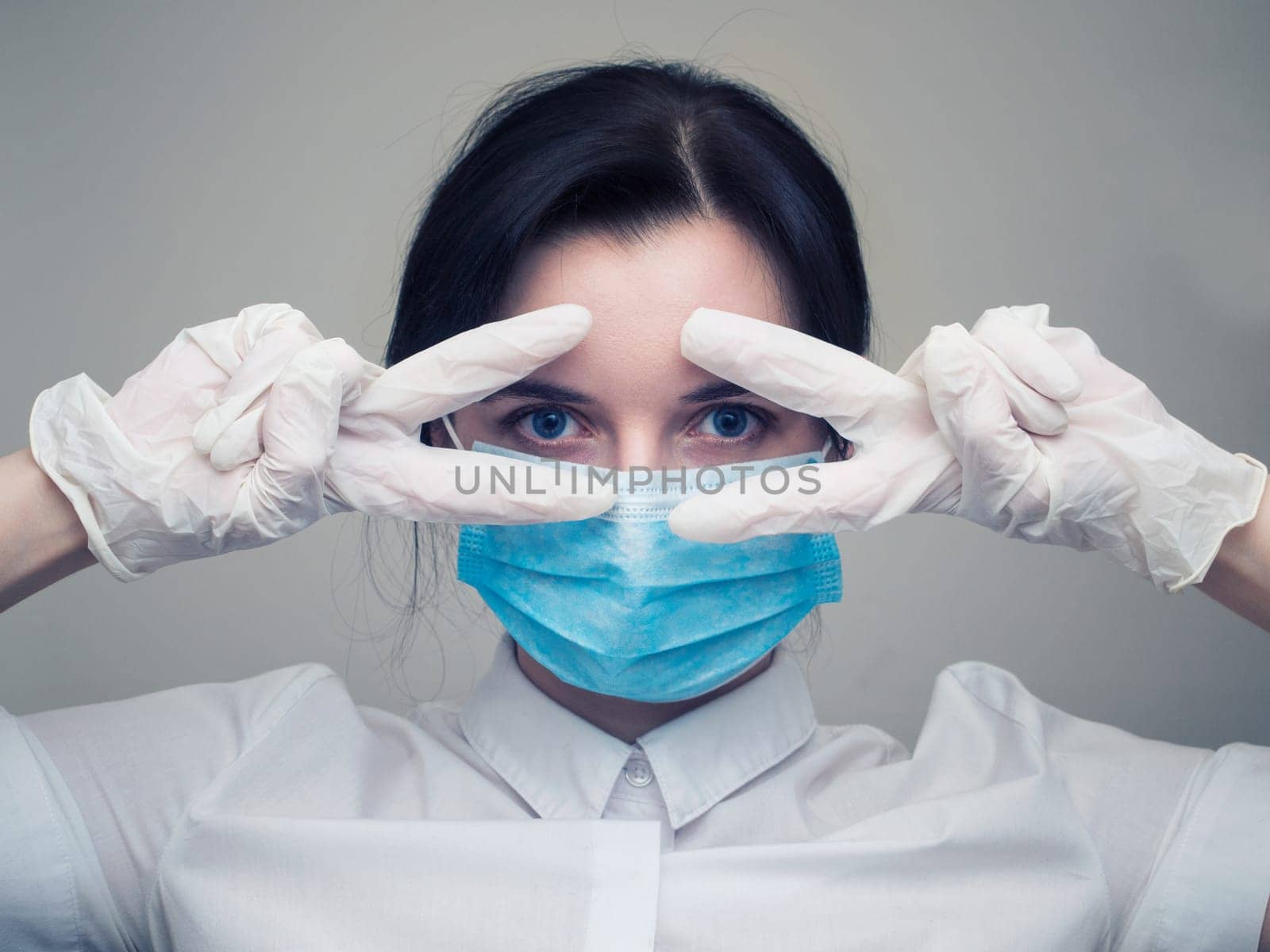 Young woman patient in a medical mask puts on protective surgical sterile gloves on her arm, on gray background, protection against coronovirus
