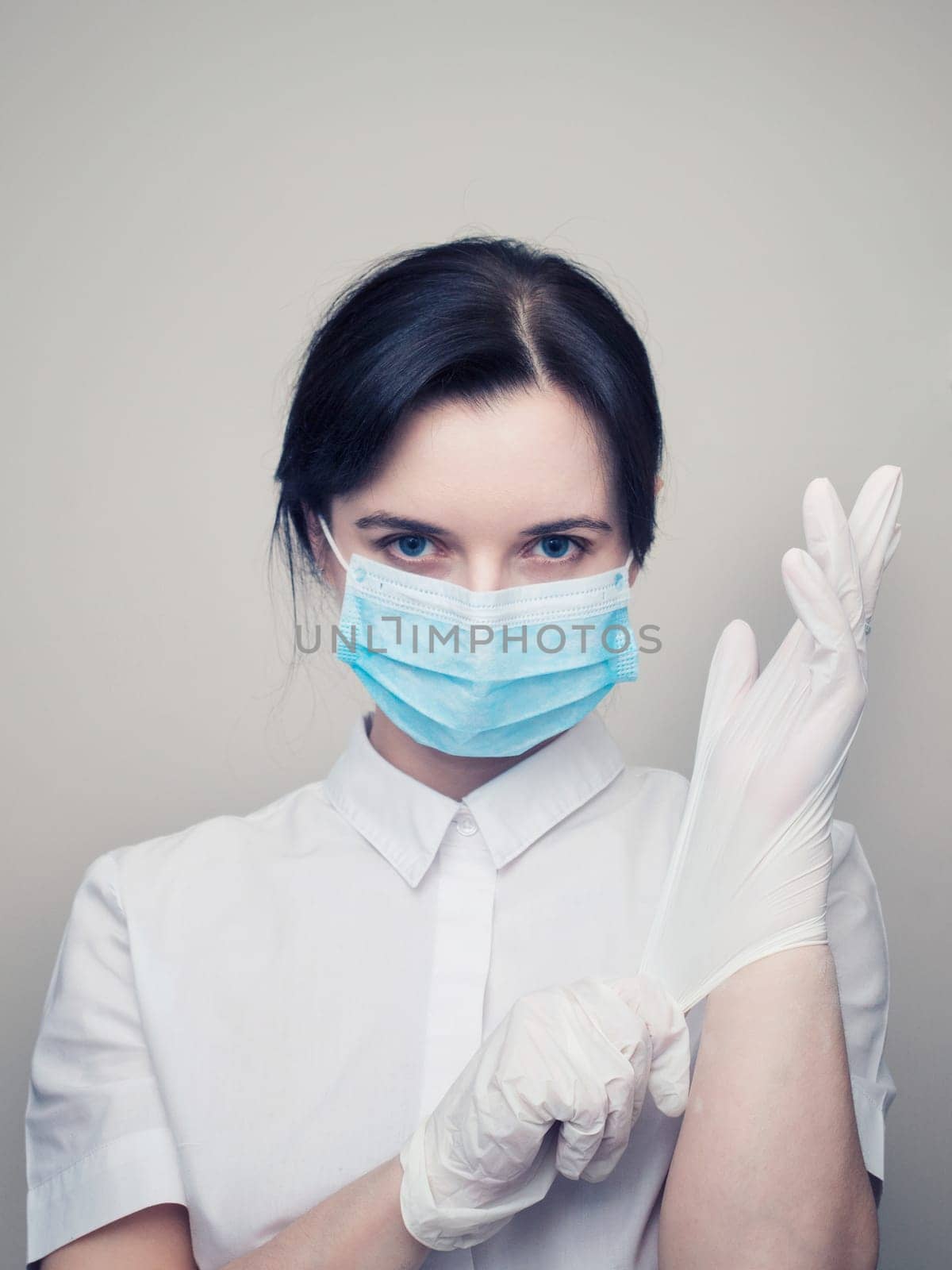 Young woman patient in a medical mask puts on protective surgical sterile gloves on her arm, on gray background, protection against coronovirus. by Andre1ns