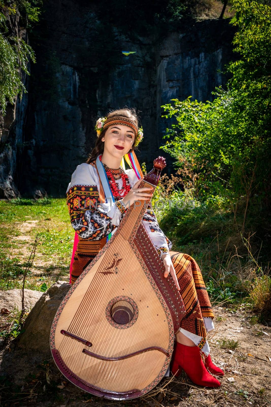 Ukrainian musician in authentic national dress sits by a rock. Ukrainian woman with a bandura musical instrument before performing folk music
