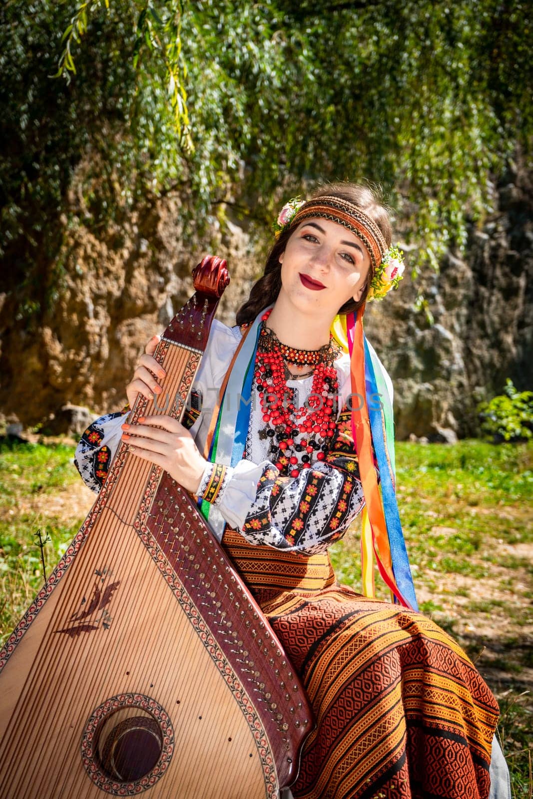 Ukrainian musician in authentic national dress sits by a rock. Ukrainian woman with a bandura musical instrument before performing folk music