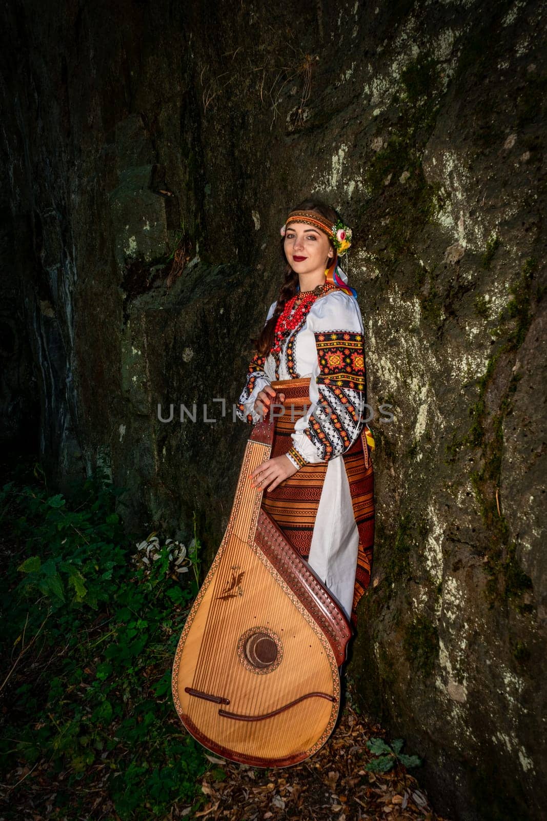 A Ukrainian woman in an embroidered national dress stands near a rock. A young woman with the Ukrainian national instrument - the bandura