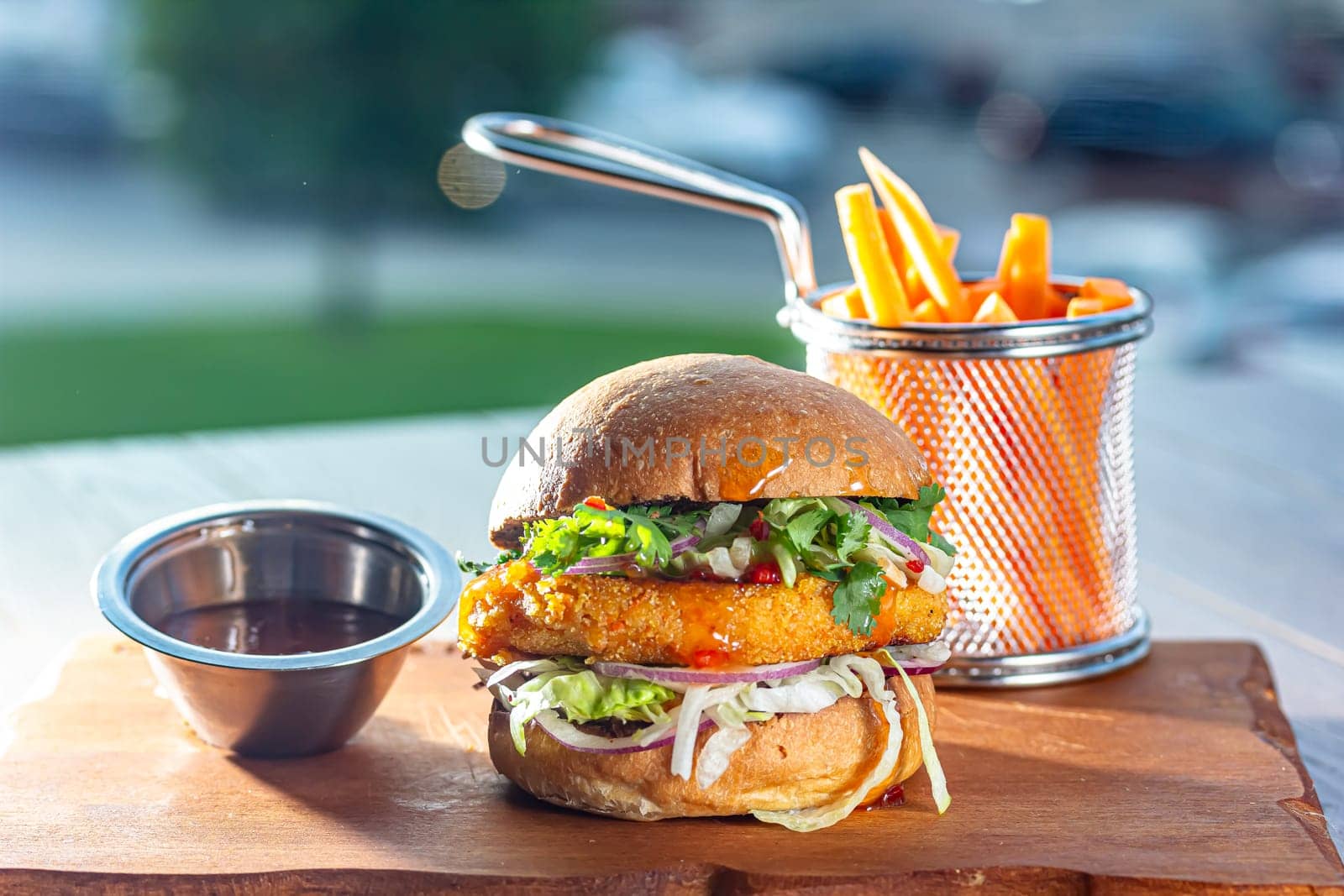 Hamburger with chicken cutlet, fresh tomatoes, red onion, lettuce and tomato sauce. Next to the burger is a metal mold with fries, a metal mold with chili sauce. Burger, fries and sauce are on a brown.
