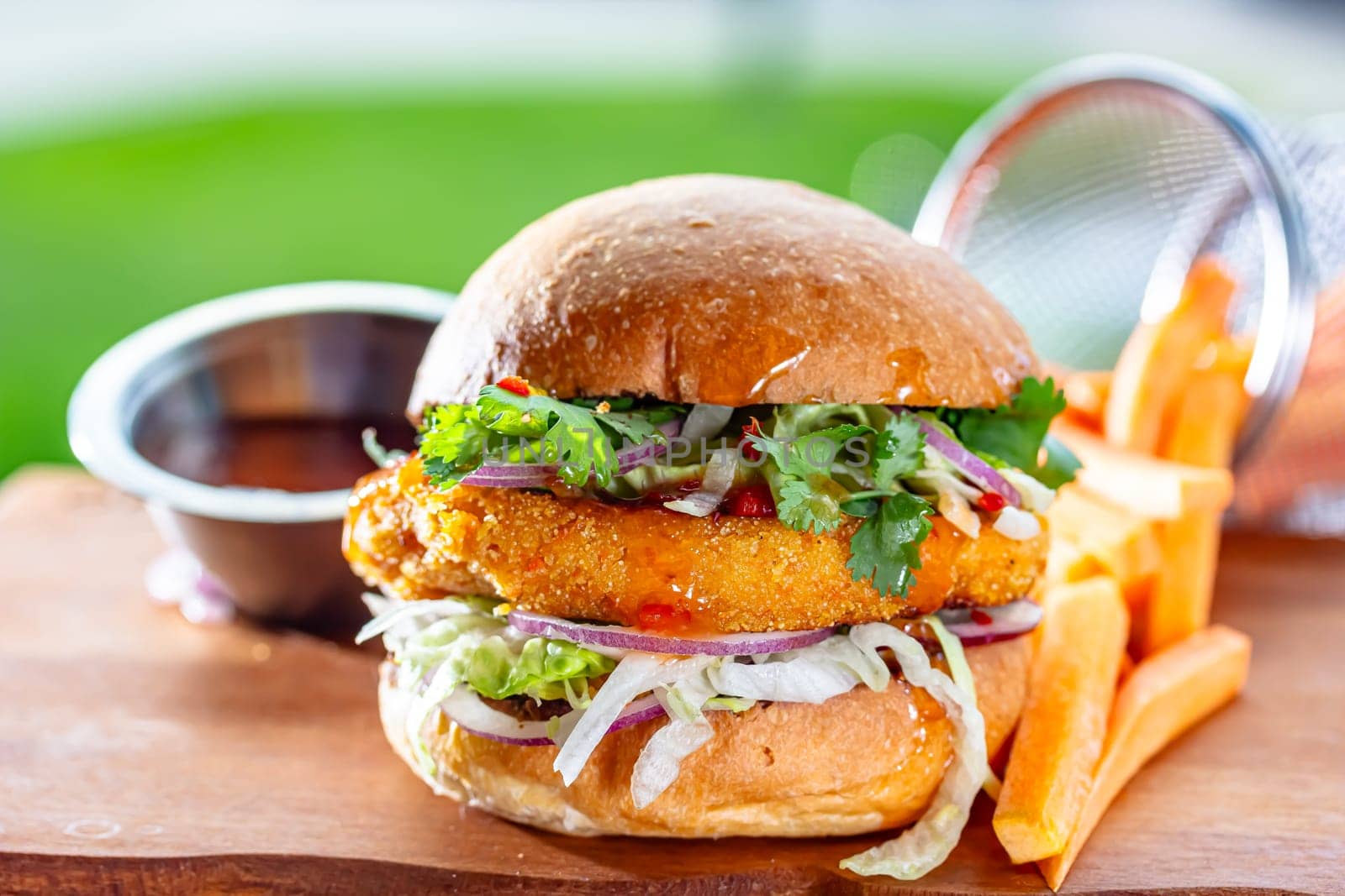 Hamburger with chicken cutlet, fresh tomatoes, red onion, lettuce and tomato sauce. Next to the burger is a metal mold with fries, a metal mold with chili sauce. Burger, fries and sauce are on a brown.