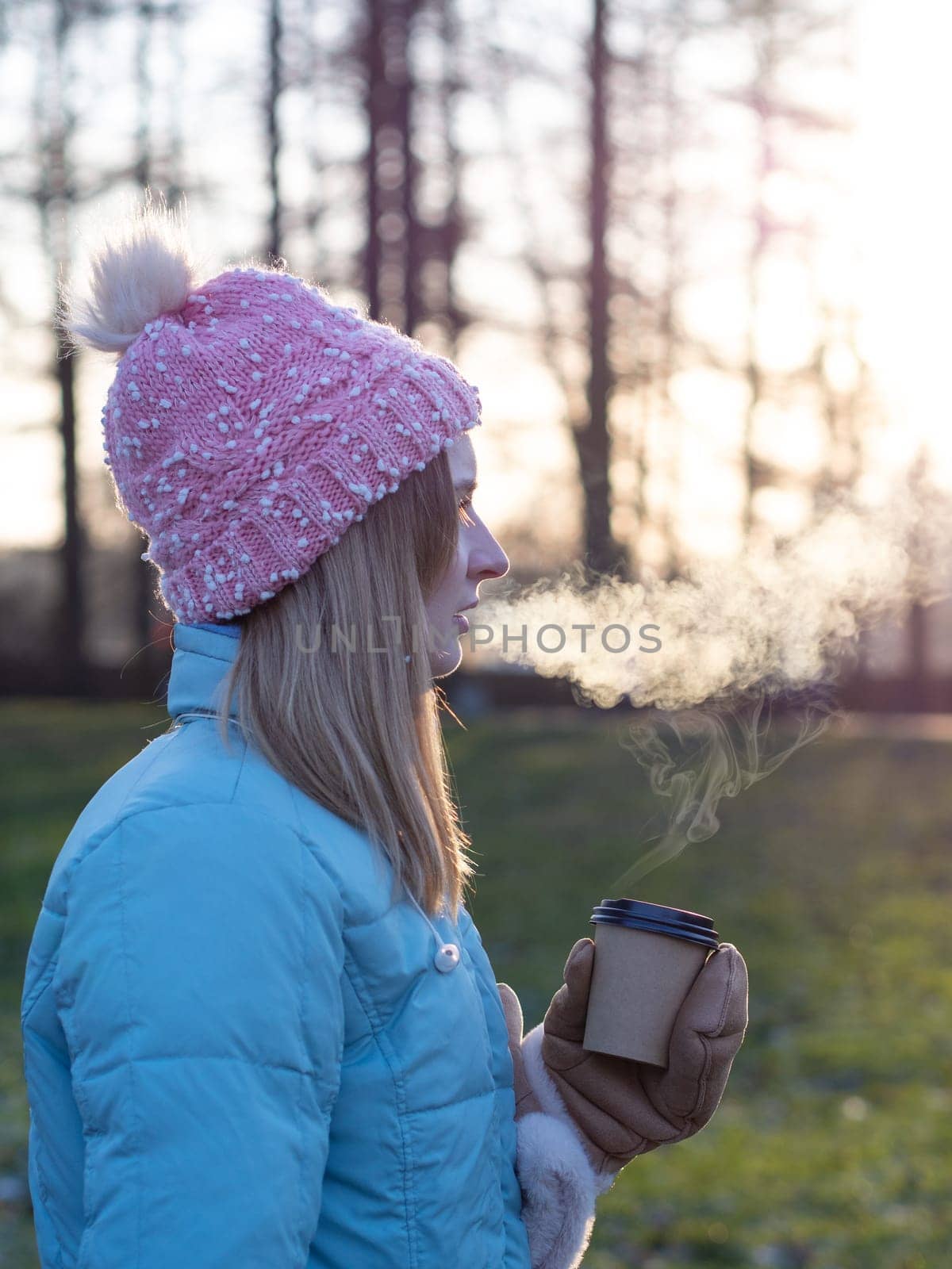 A beautiful cute girl in a hat and a down jacket holds a paper cup with hot coffee or tea. Frosty sunny morning Winter season by Andre1ns