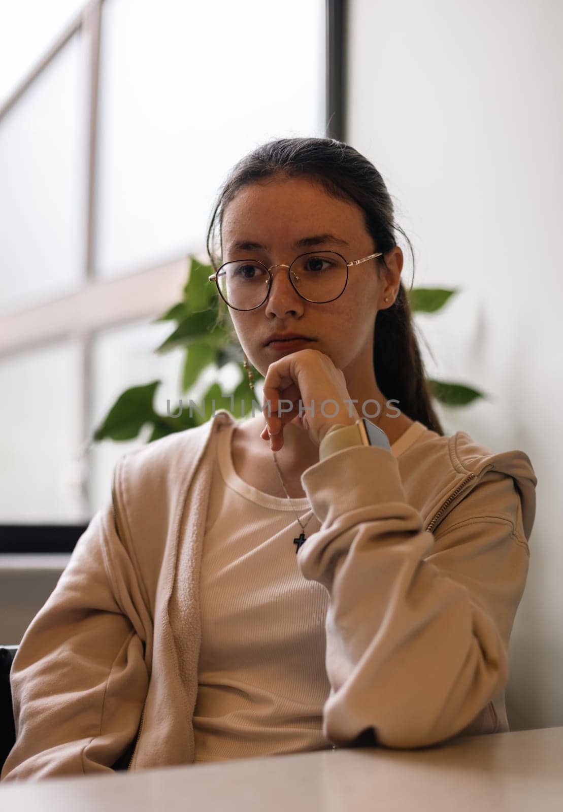 Portrait of one beautiful caucasian brunette teenager girl in glasses in beige clothes sits in a cafe against the background of a panoramic window looking to the side and holding her chin with her hand, side view close-up.