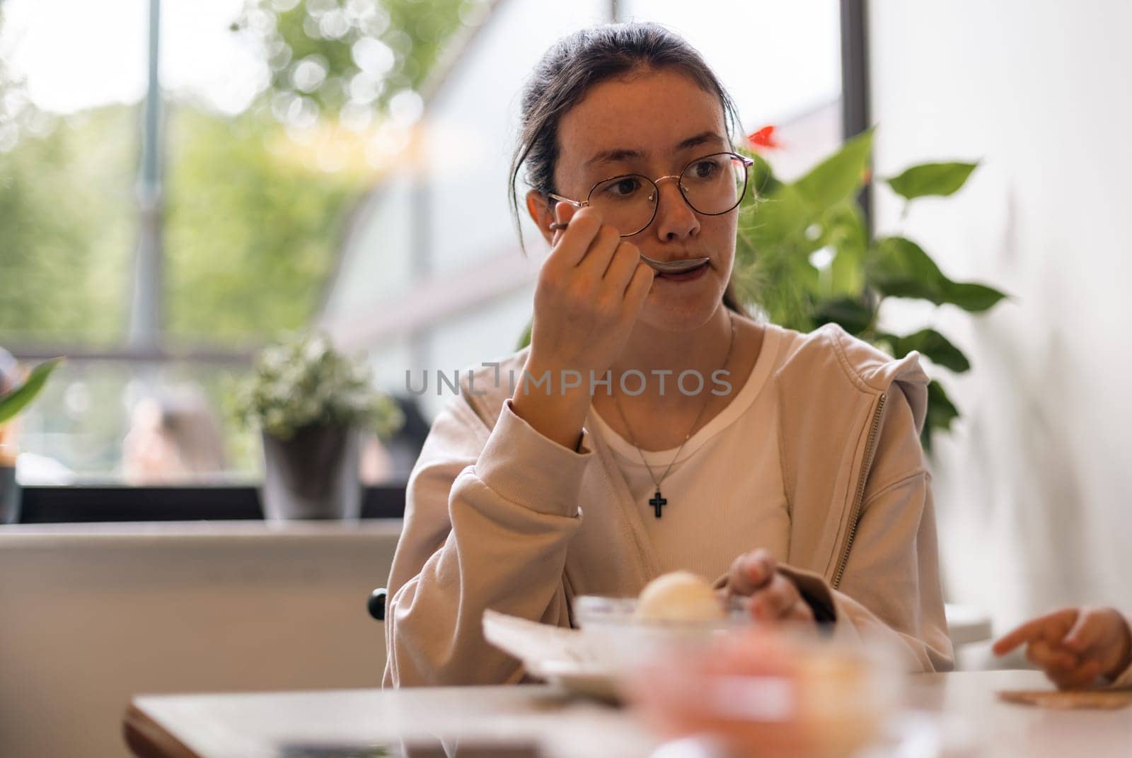 Portrait of one beautiful caucasian sad teenage girl eating ice cream and looking away while sitting on a sofa in a cafe against the background of a panoramic window, close-up side view.