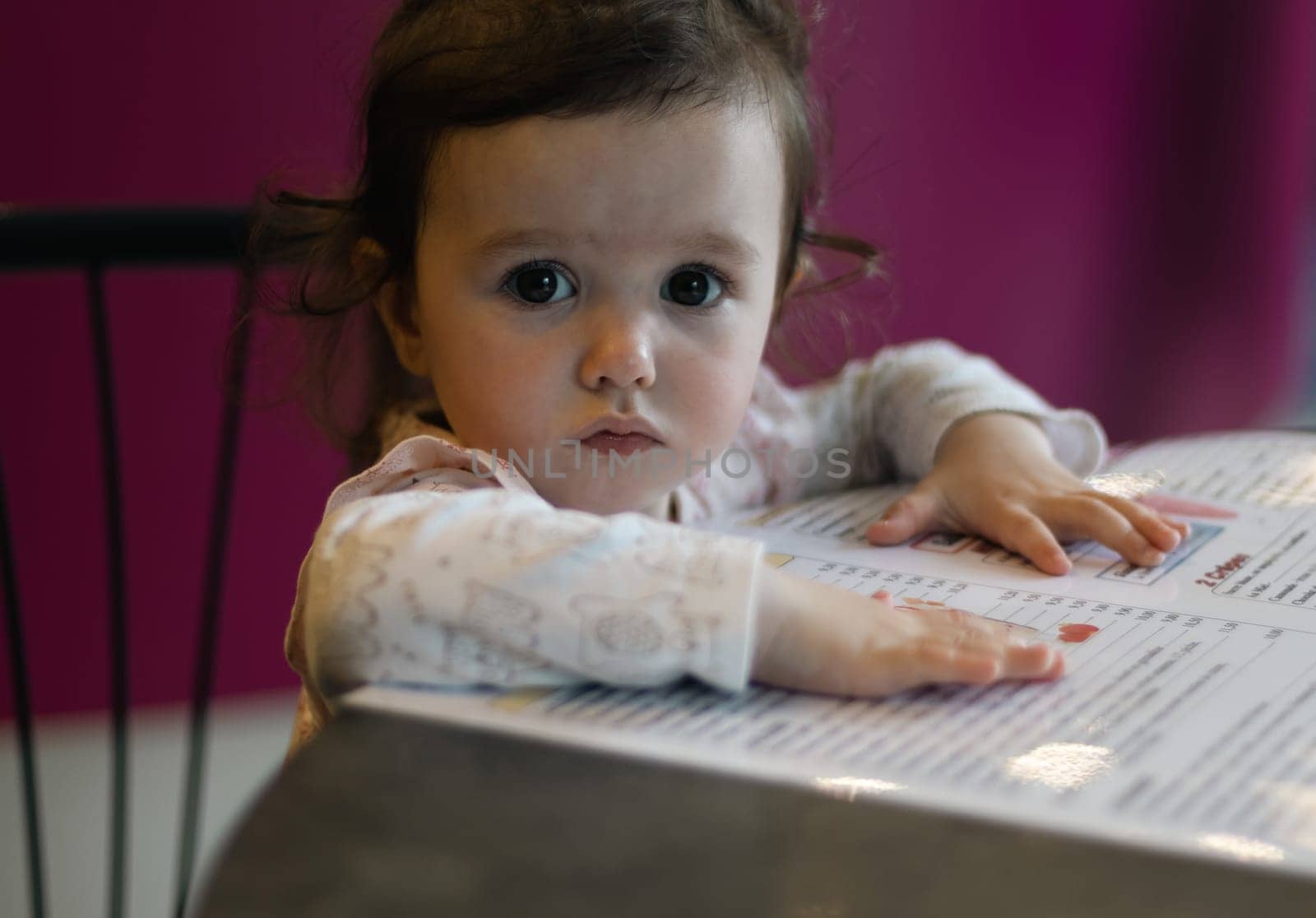 Portrait of one beautiful caucasian baby girl sitting in a cafe at a table, choosing a dessert on the menu and looking at the camera,