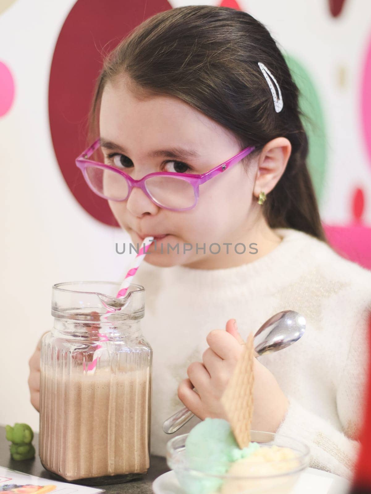 Portrait of a beautiful Caucasian brunette girl in glasses sitting at a table with ice cream and drinking a chocolate milkshake from a straw from a glass mug in a cafe on a blurred abstract background, close-up side view with depth of field.