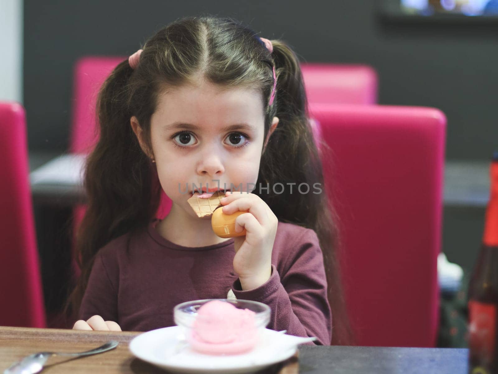 Portrait of a beautiful Caucasian brunette girl with big brown eyes and two ponytails on her head sitting at a table and eating an ice cream waffle in a cafe on a blurred background, close-up side view with depth of field.