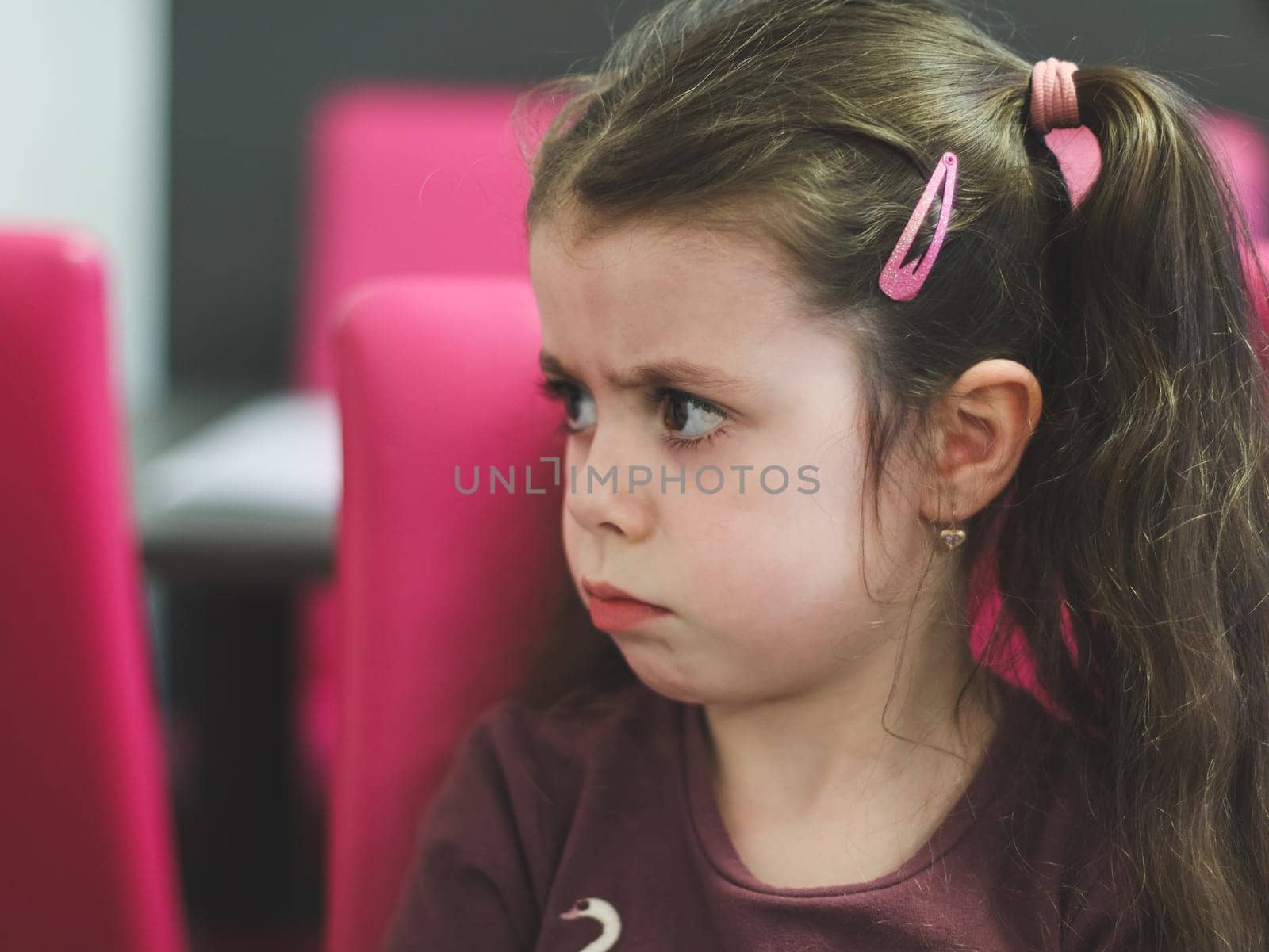 Portrait of one beautiful Caucasian girl with big brown eyes sitting at a table in a cafe with an offended emotion on her face and pouty lips, on a blurred background, close-up side view with depth of field.