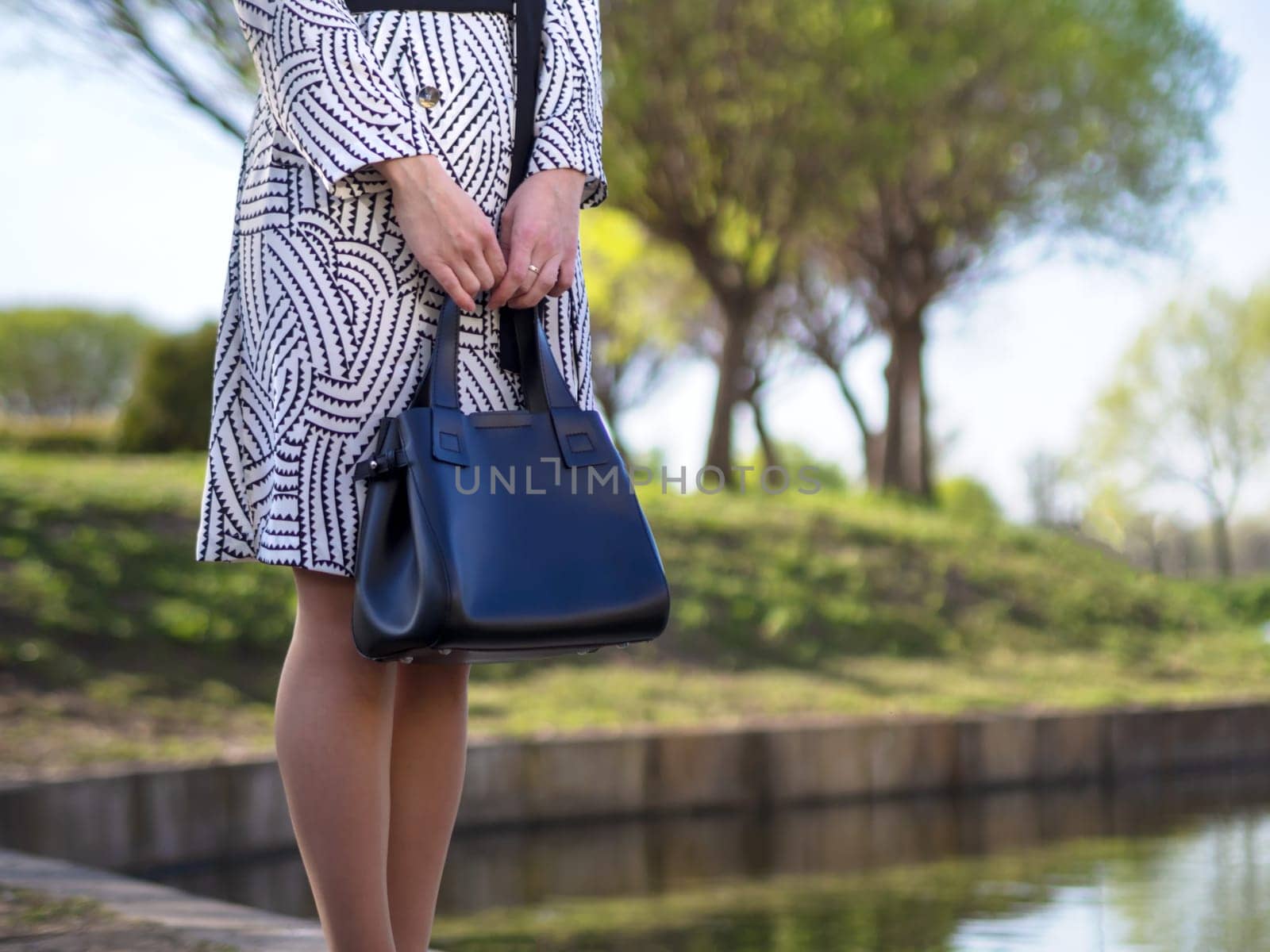 stylish European young woman in a raincoat, tights, shoes with heels, with a black leather bag in her hands in a park near the lake