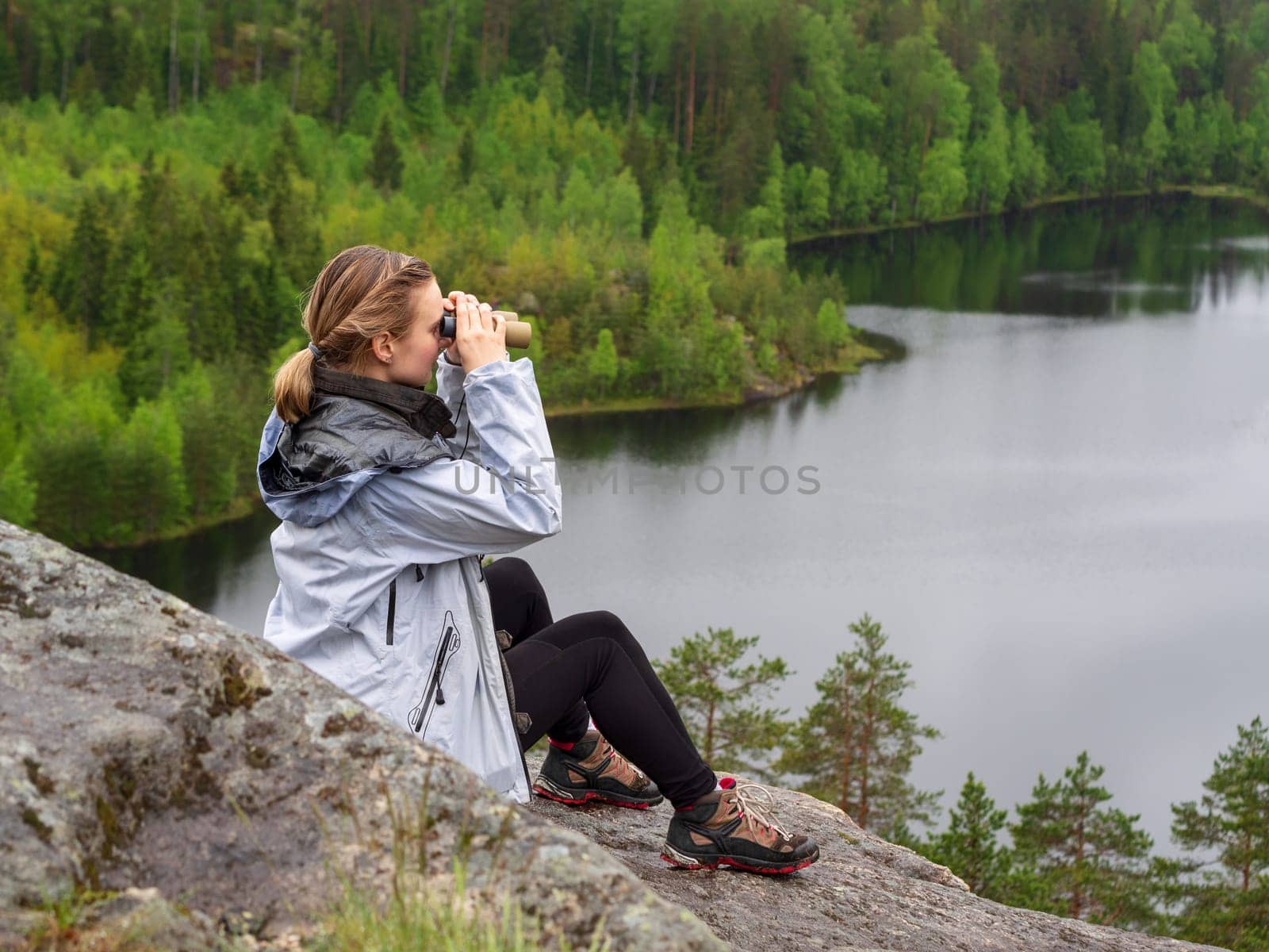 woman looks through binoculars. tourist girl on a rock in a forest near a lake