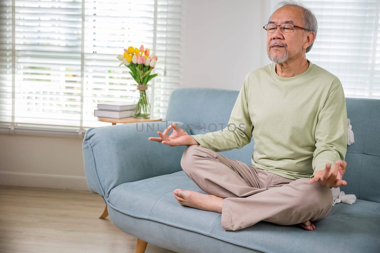 Lifestyle senior man sitting on sofa in living room holding hands in mudra practicing home yoga in lotus pose, Elderly relaxed man do physical exercises following healthy on stress free weekend