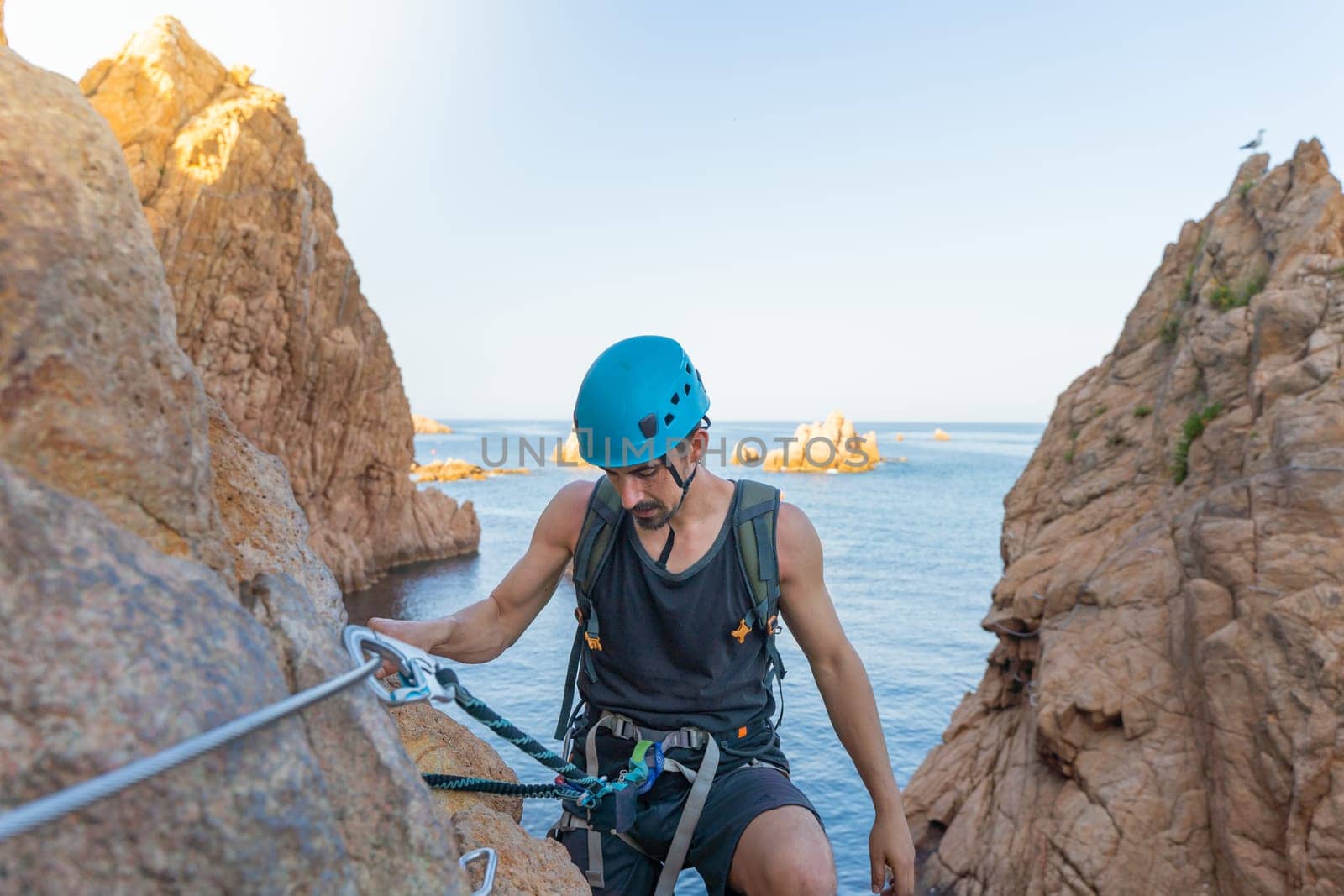 Young male climber strong success hiking on rocks over the sea with rope and helmet. by PaulCarr