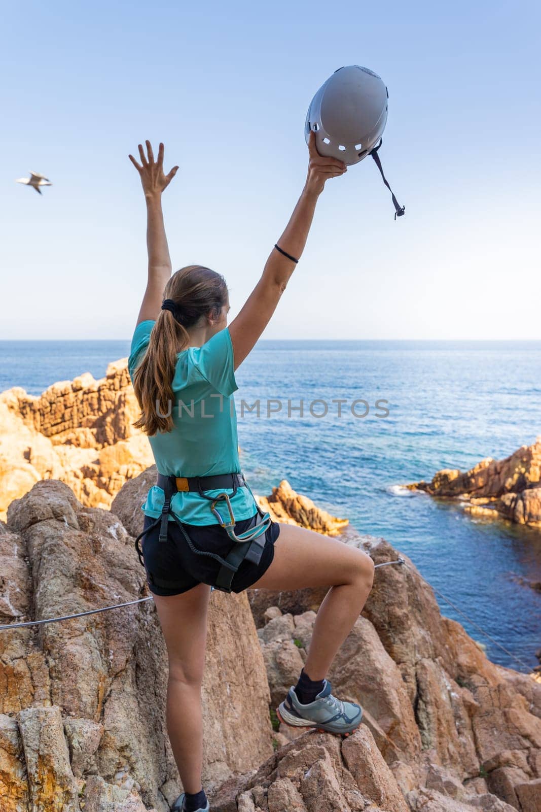 Pretty female climber celebrating strong success with arms raised on rocks over the sea with rope and helmet. High quality photo