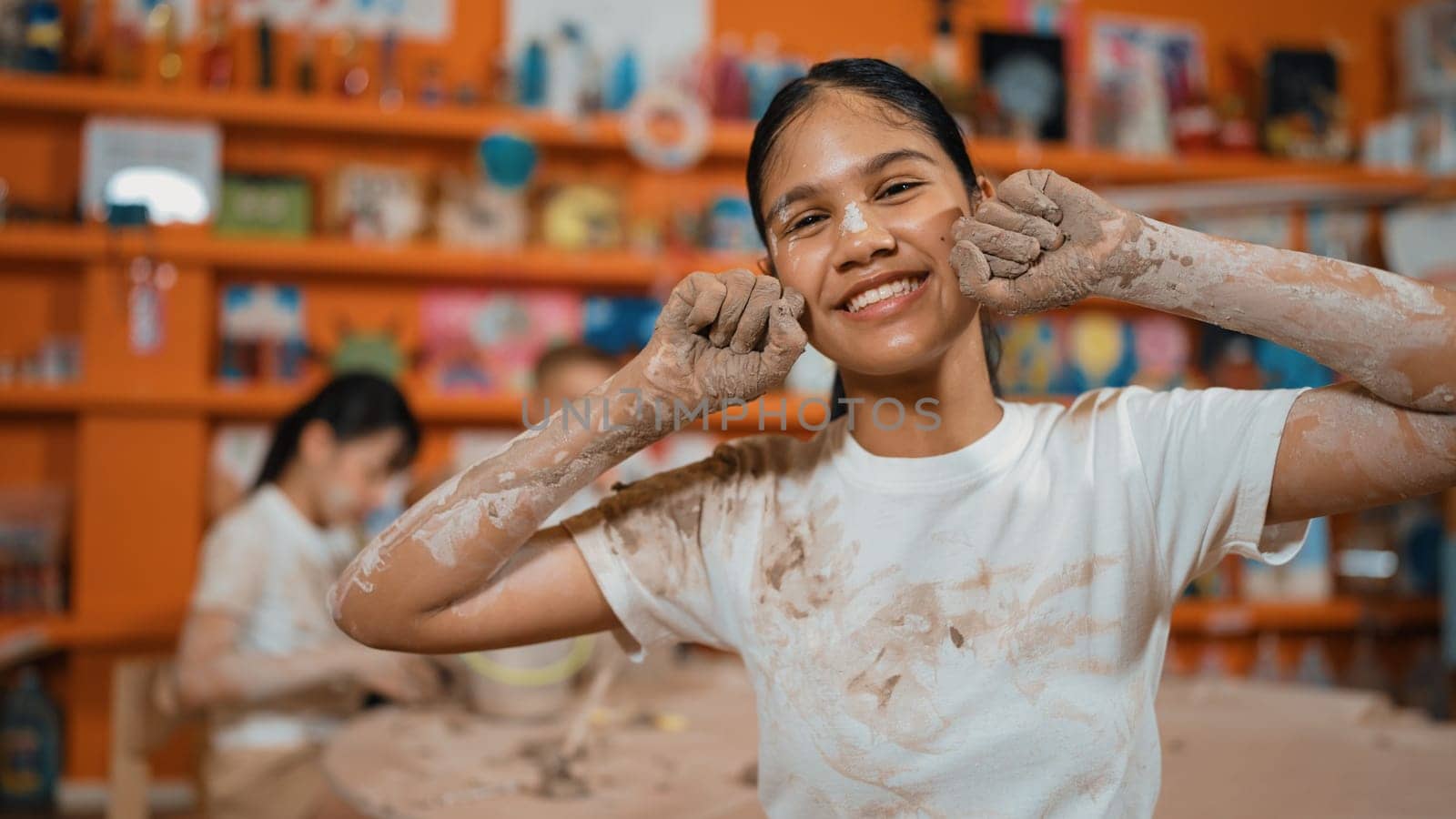 Happy caucasian girl pose at camera while diverse children modeling clay behind. Cute student wearing dirty shirt while looking at camera at workshop in art lesson. Blurring background. Edification.